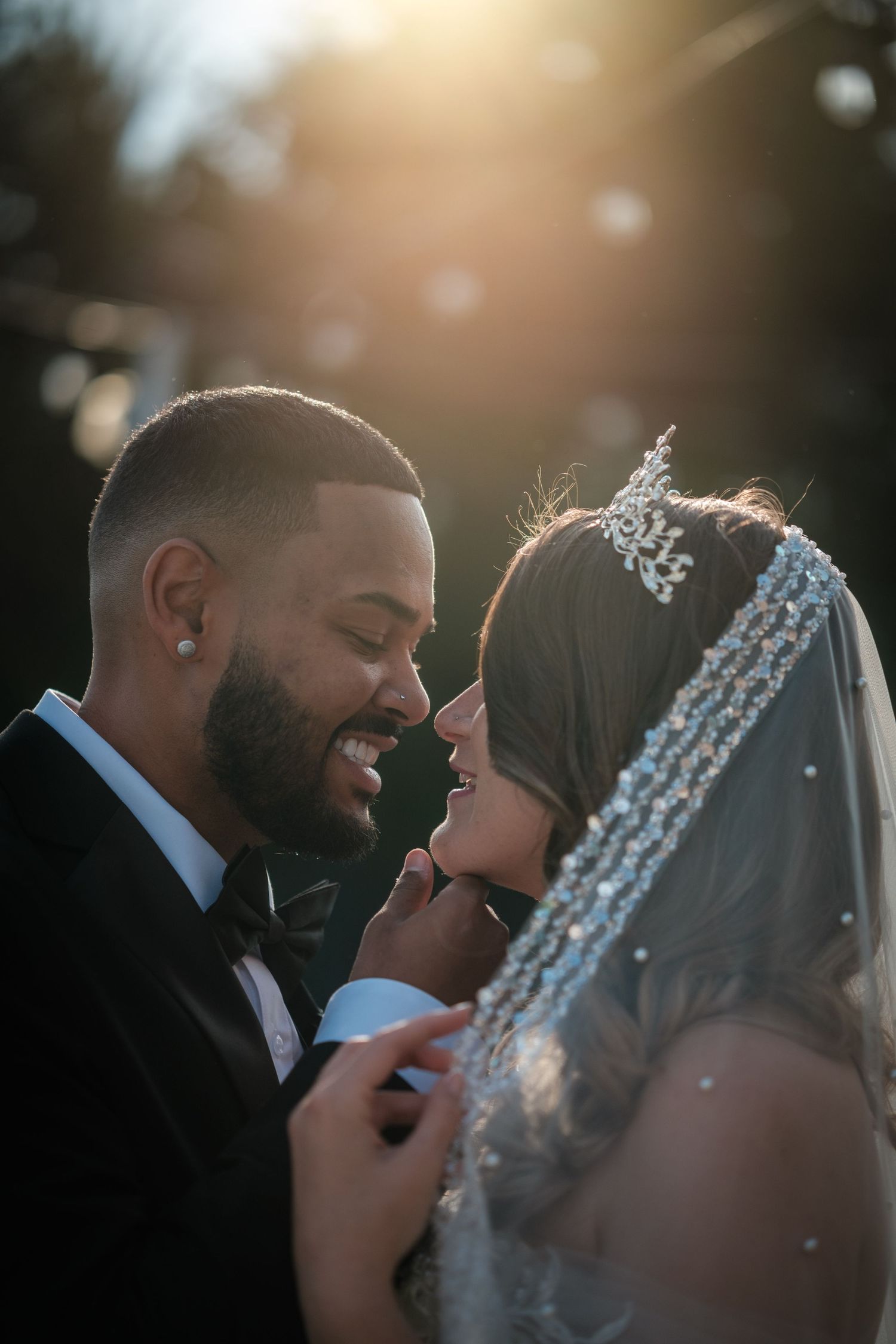 A bride and groom share a romantic moment outdoors, bathed in golden sunlight, as she wears a beaded crown and veil.