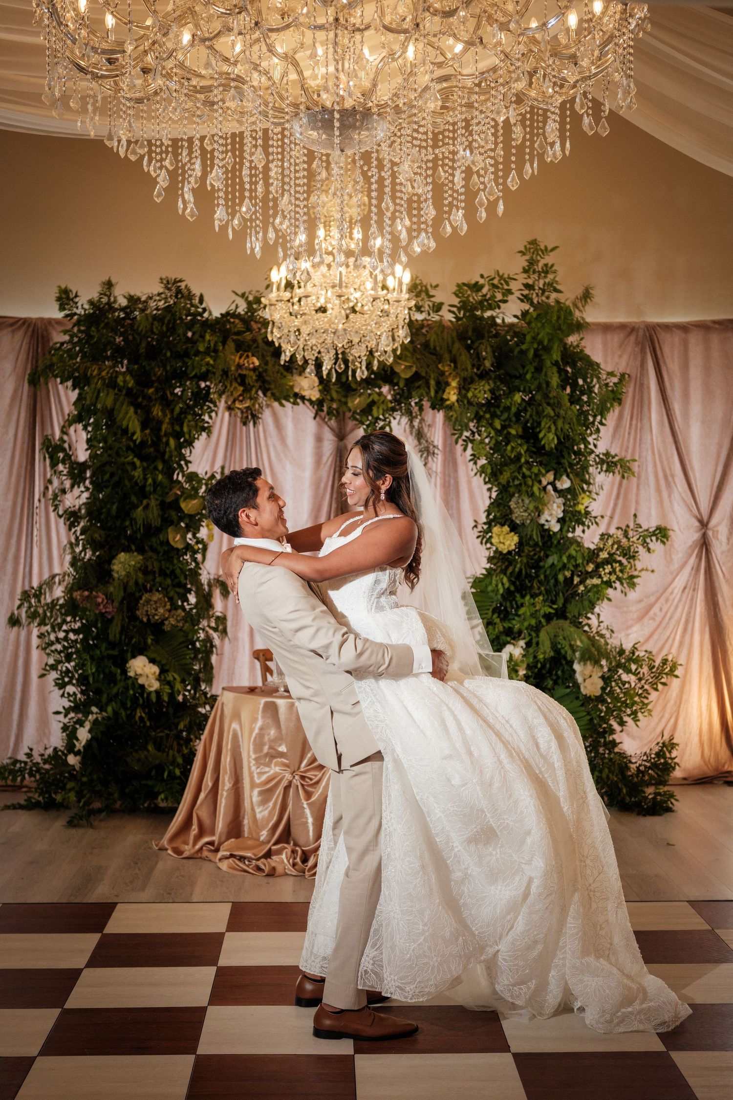 Groom lifts bride in white lace gown under crystal chandelier, posing before lush greenery floral arch at elegant wedding reception.