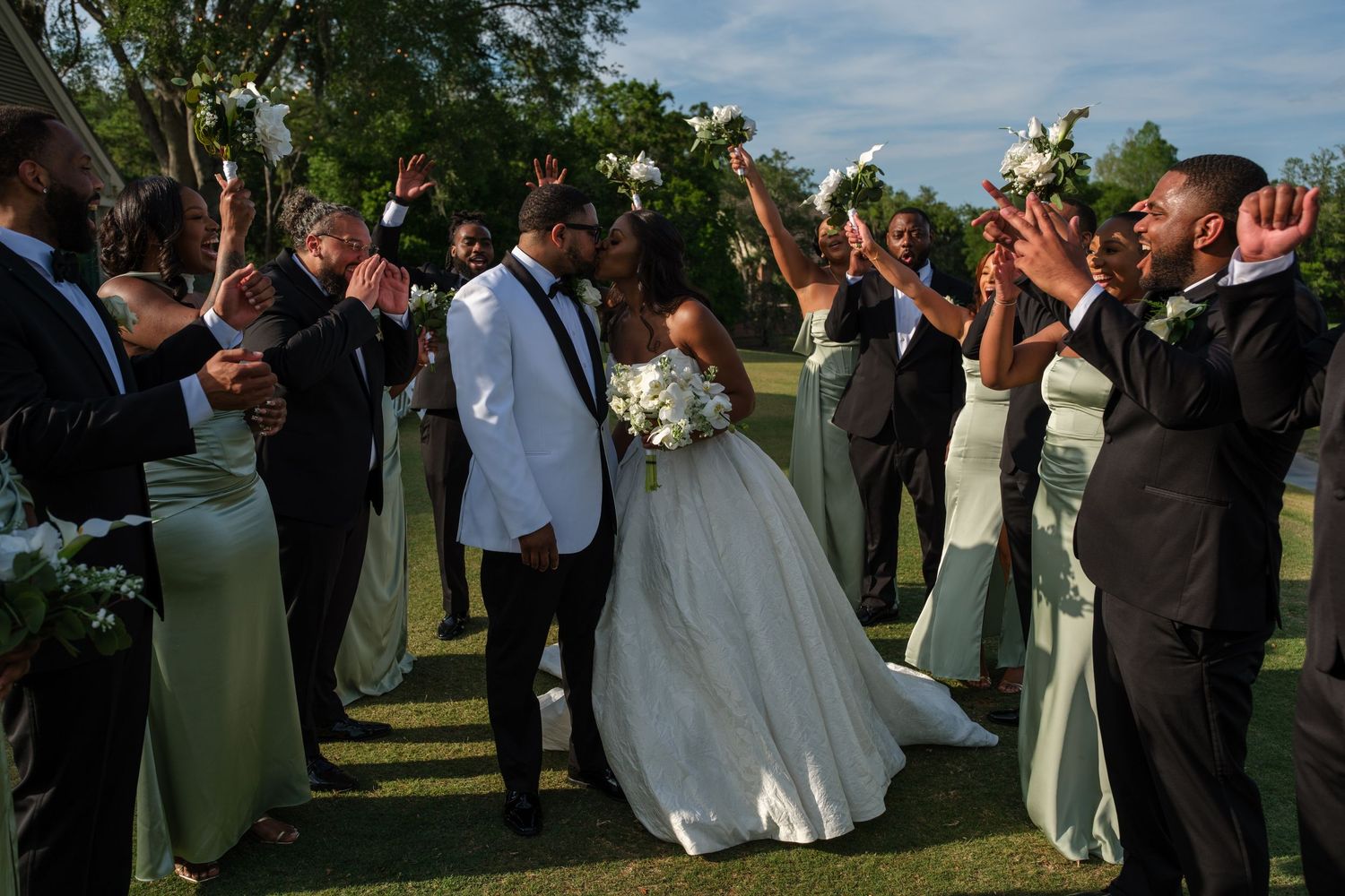 Bride and groom kiss surrounded by cheering wedding party in sage green and black attire on a sunny outdoor lawn.