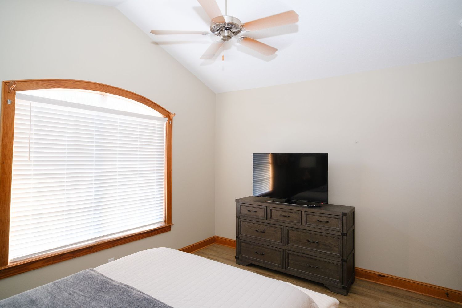 Modern bedroom interior with arched window, ceiling fan, and dark wood dresser with mounted TV on top.