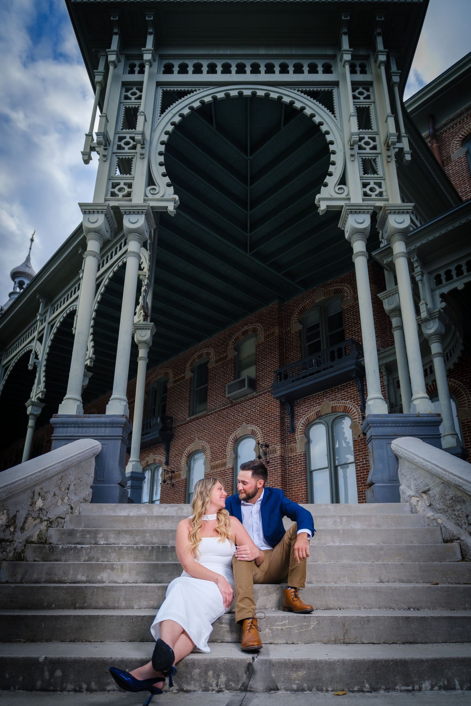 A couple sits together on grand stone steps of a Victorian-style building at the University of Tampa during their engagement photo session