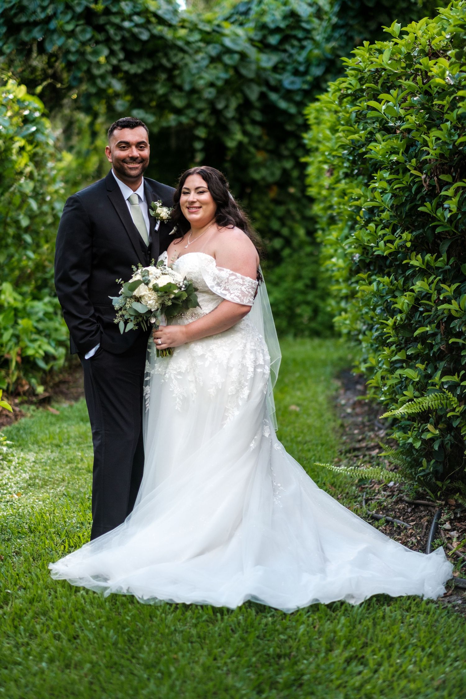 Wedding couple poses in a lush garden setting with flowing white gown and classic black tuxedo.