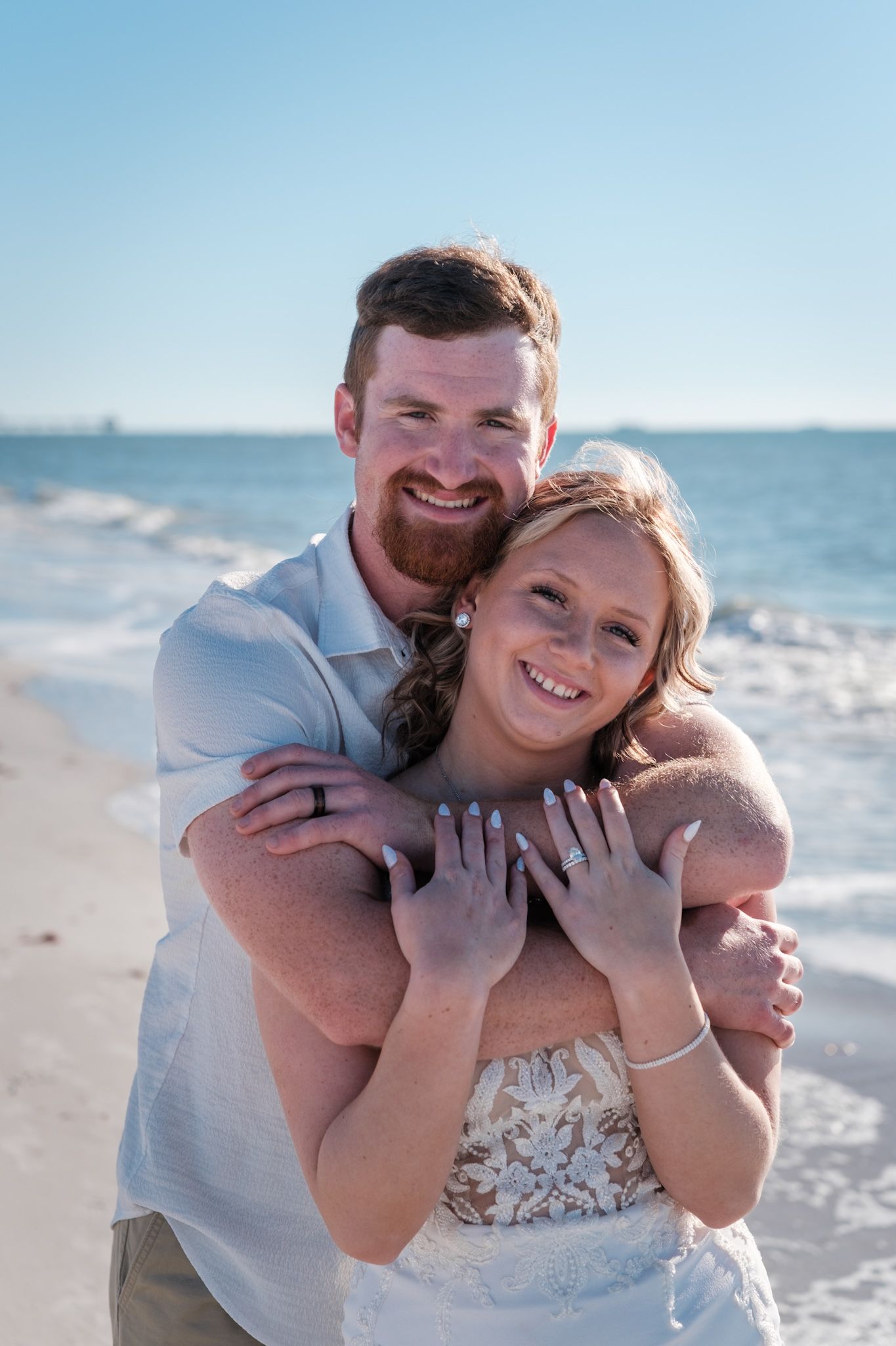 A joyful couple embraces on a sunny beach, with waves and blue sky in the background.