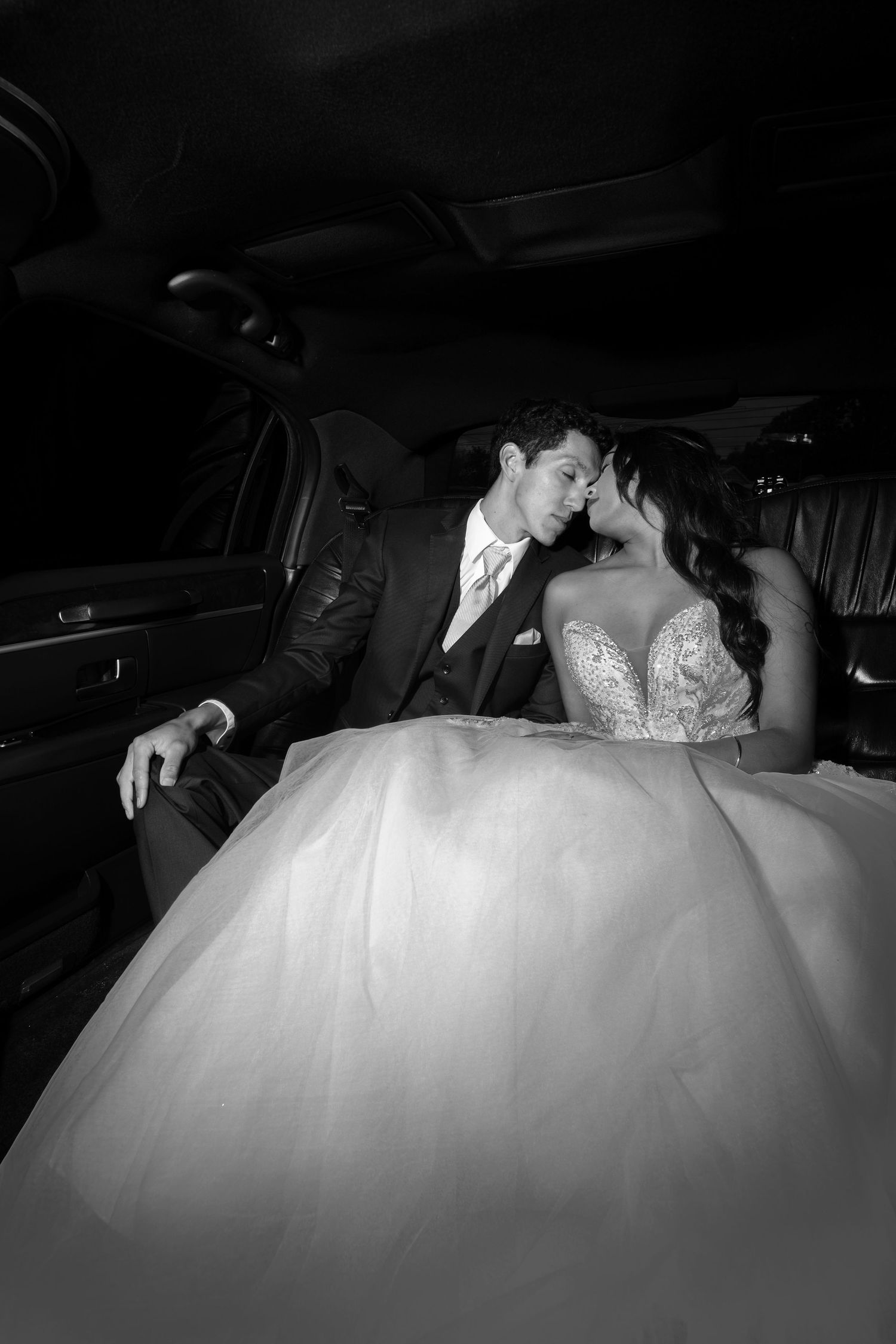A couple in wedding attire sitting together in the backseat of a dark limousine.
