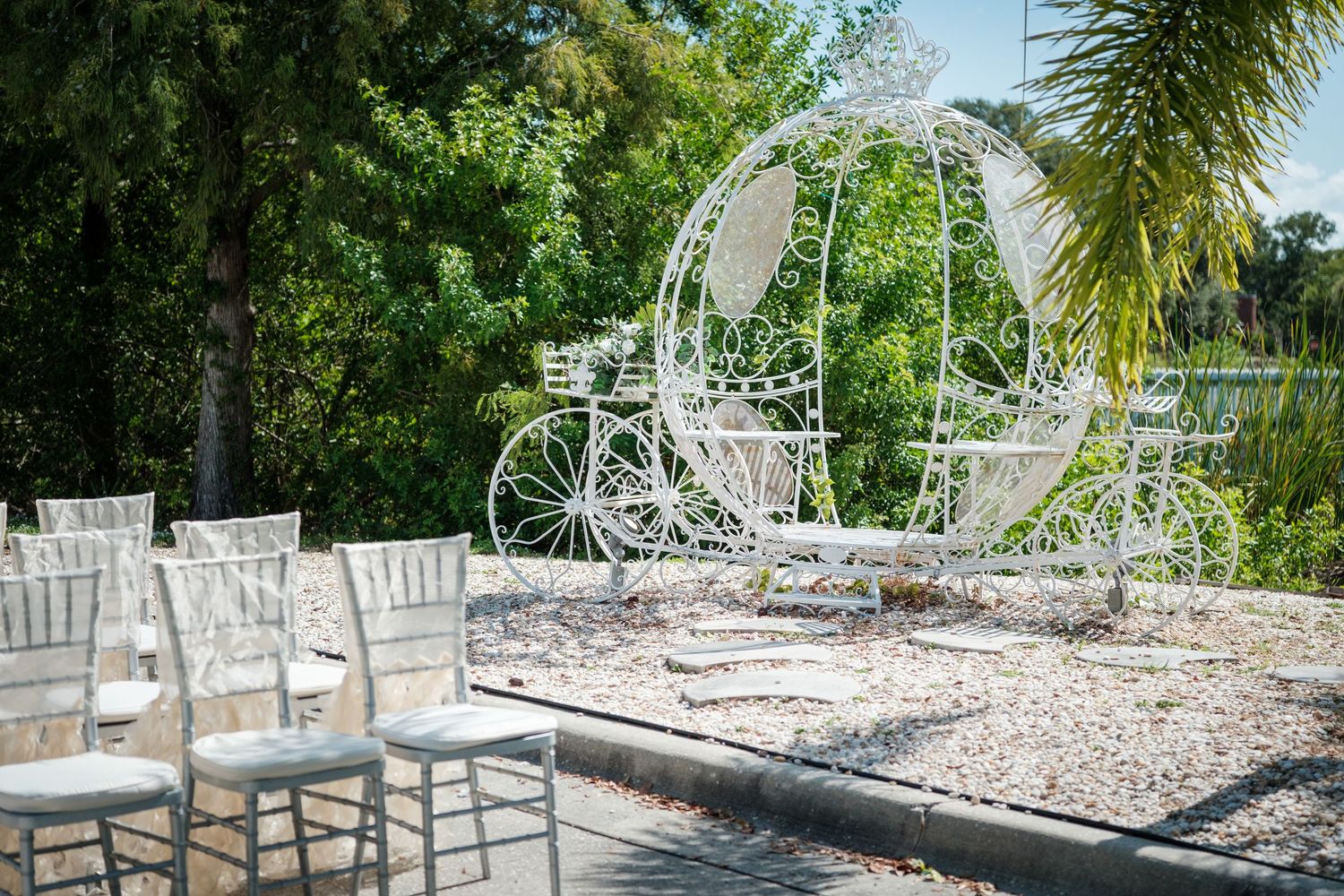 White ornate Cinderella carriage wedding decoration outdoors with chiavari chairs on a patio surrounded by lush greenery.