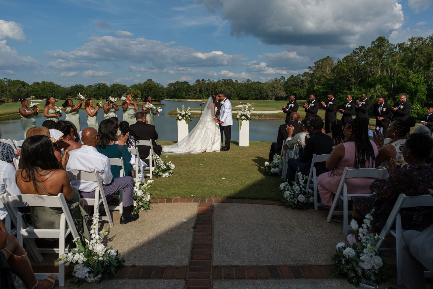 Outdoor wedding ceremony by a lake with bride and groom at altar, guests seated in white chairs, lush greenery backdrop.