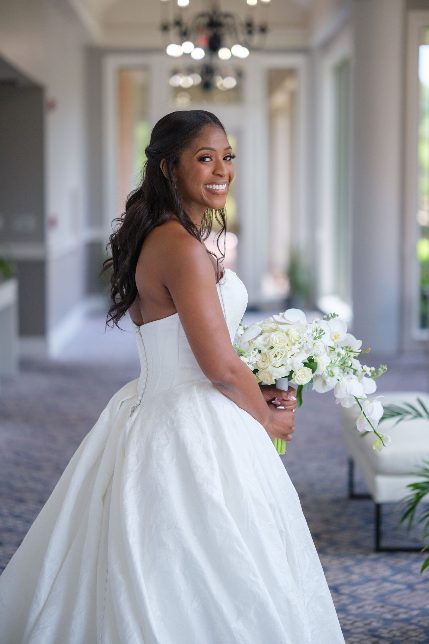 Smiling bride in strapless white ball gown holding white floral bouquet in elegant venue hallway.