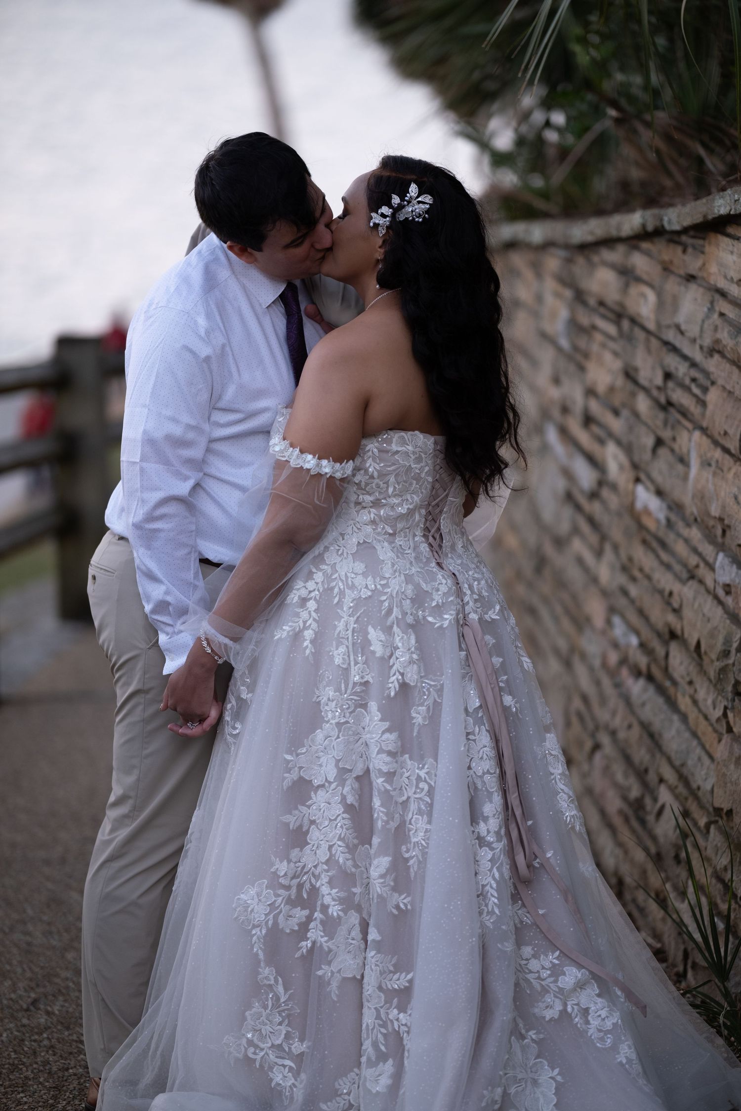 A romantic wedding couple embraces against a stone wall, with the bride wearing an elegant lace dress with floral details.