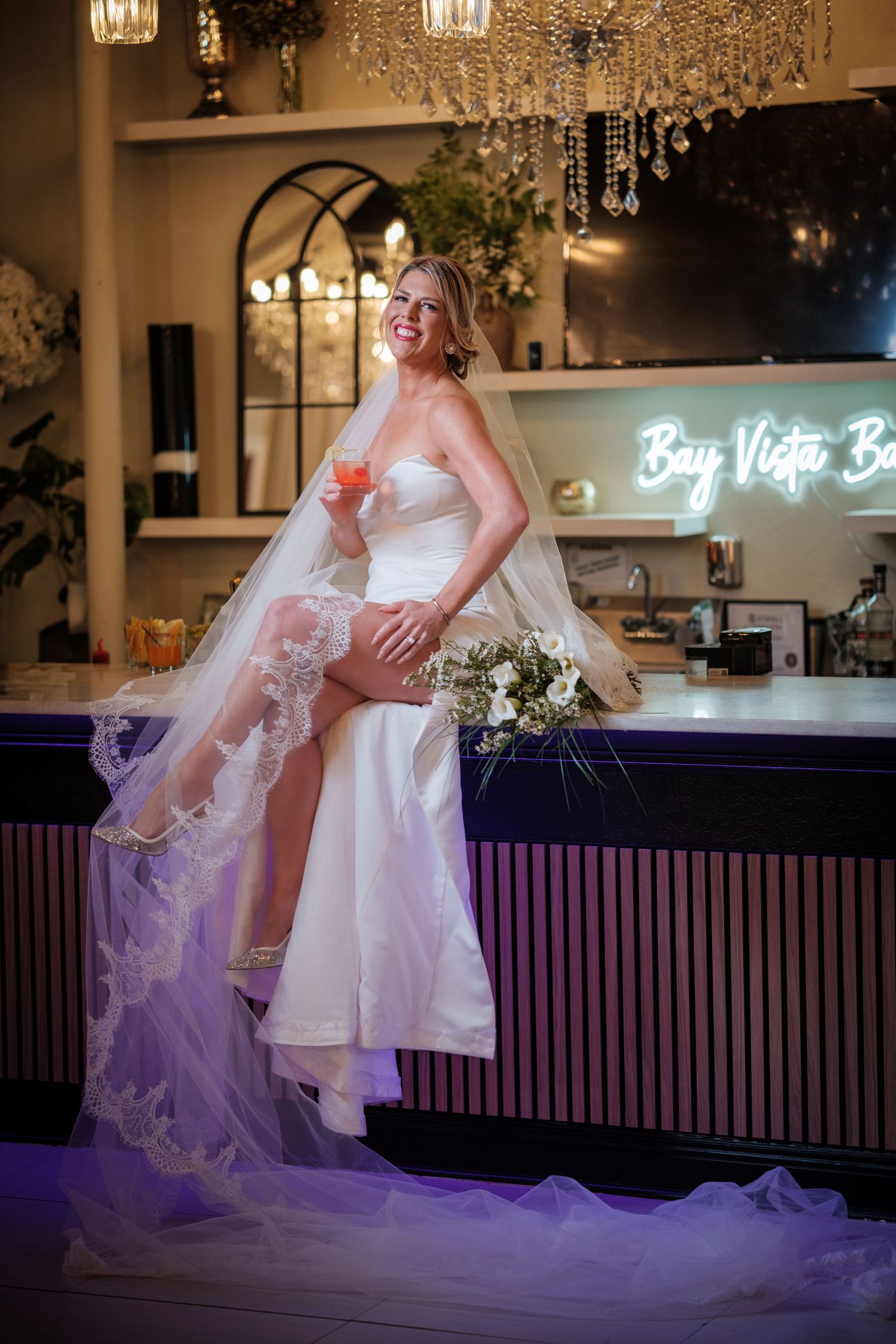 Smiling bride in white strapless gown with lace veil sits on bar counter holding bouquet and drink at Bay Vista Bar.