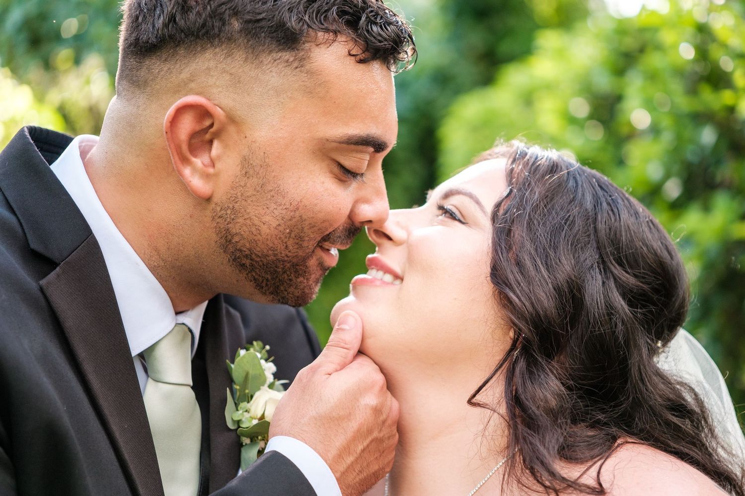 A romantic wedding couple shares a tender kiss outdoors in a garden setting with lush greenery in the background.