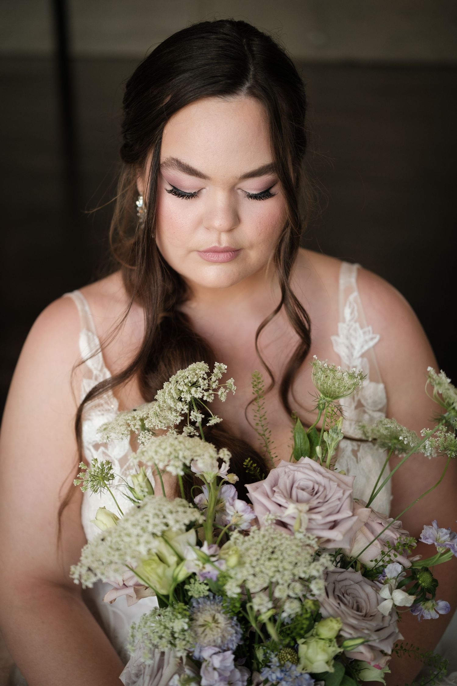 A bride in a white lace dress holds a delicate bouquet of roses and Queen Anne's lace with soft neutral colors.