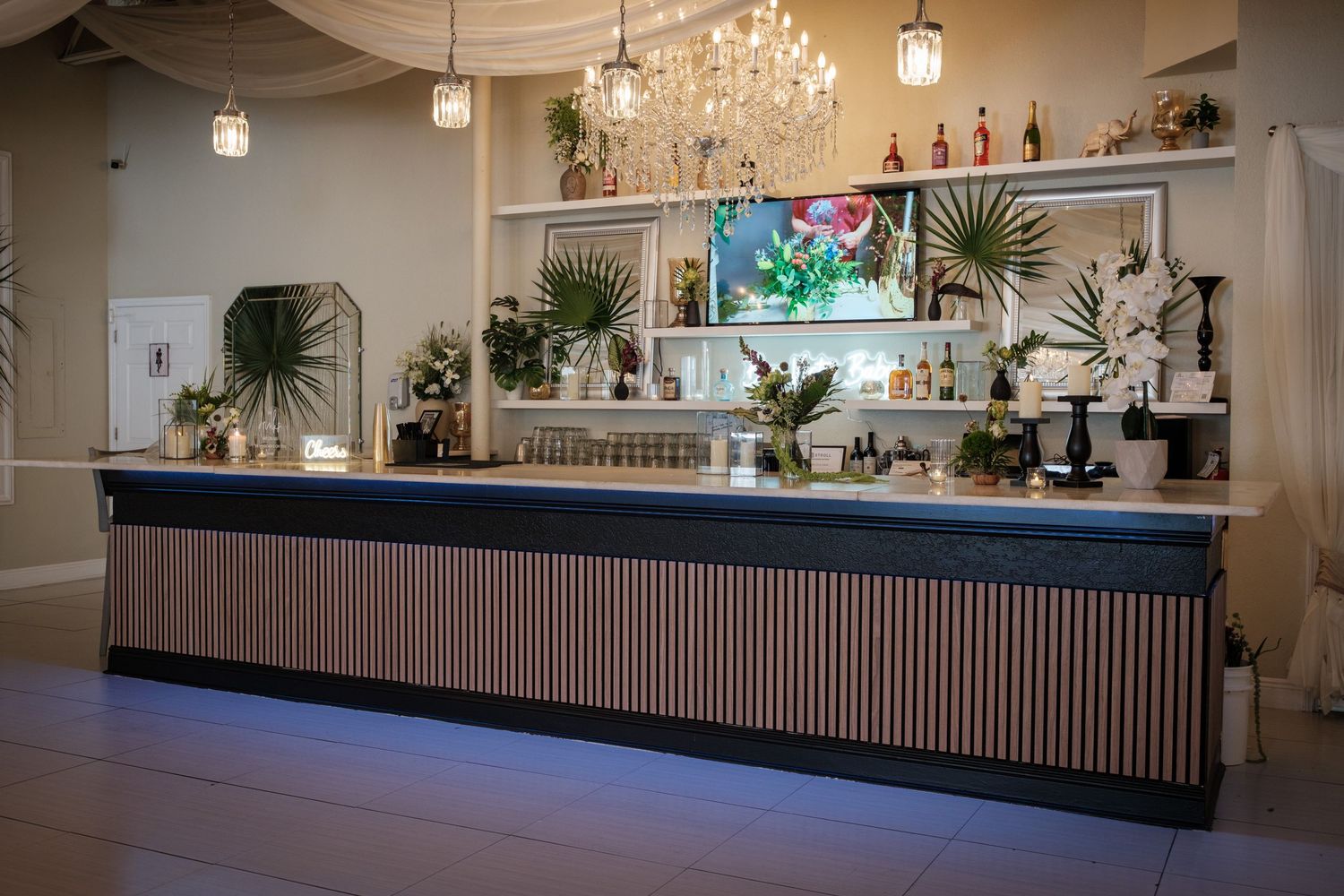 Elegant bar counter with wood paneling, lush green plants, chandelier lighting, and a well-stocked back bar display.