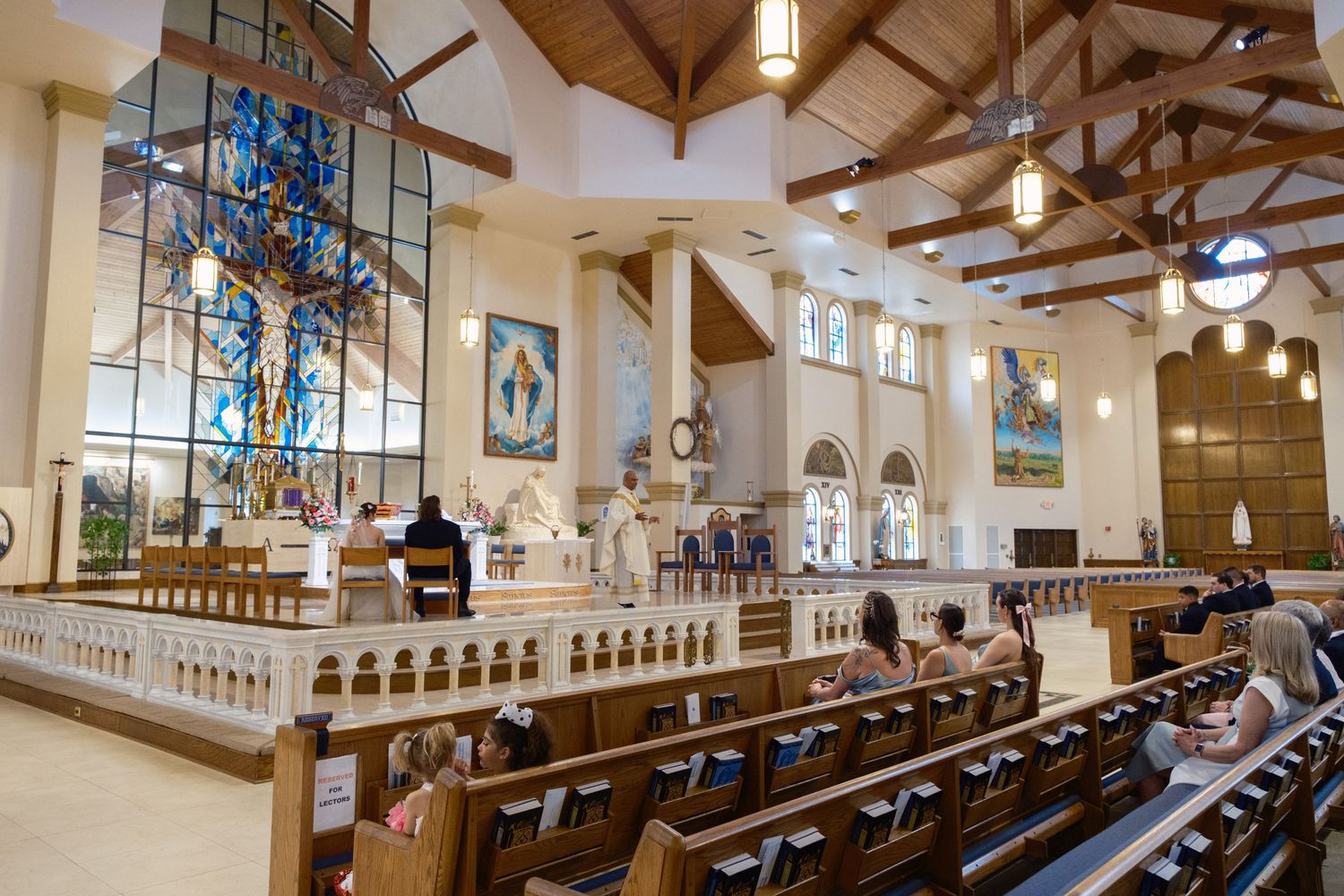 Bright Catholic church interior with stained glass windows, wooden pews, and ornate altar during a service.