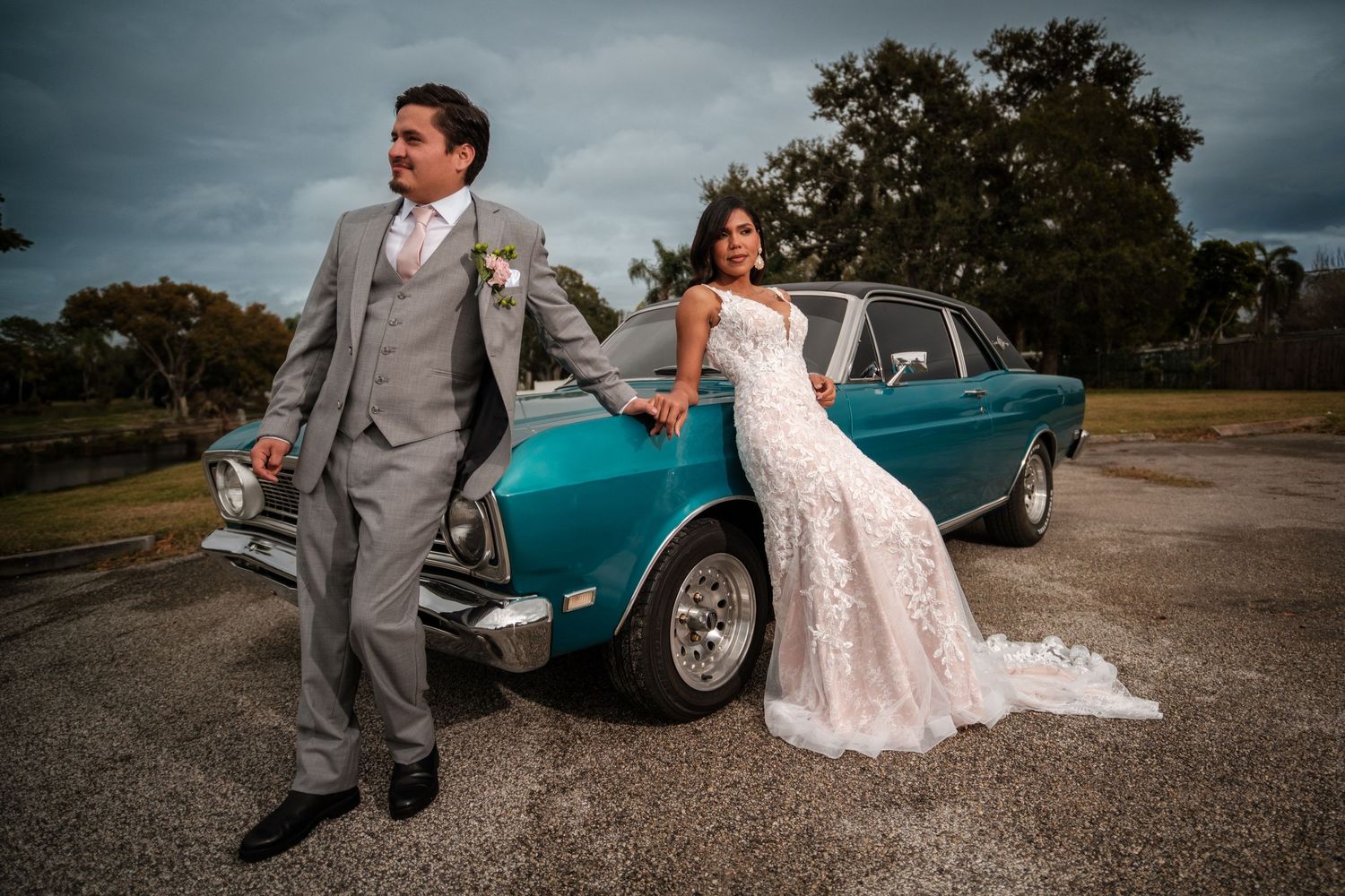 A romantic wedding couple poses together near a classic turquoise vintage car on a gravel road.