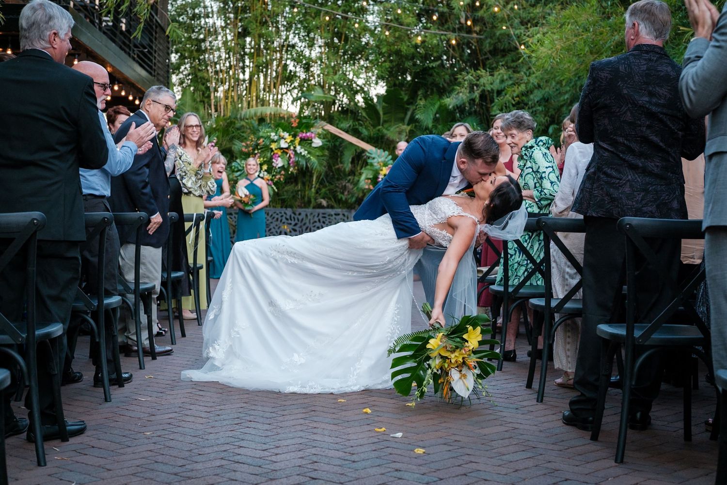 A romantic dip and kiss during an outdoor wedding ceremony surrounded by guests and lush greenery.