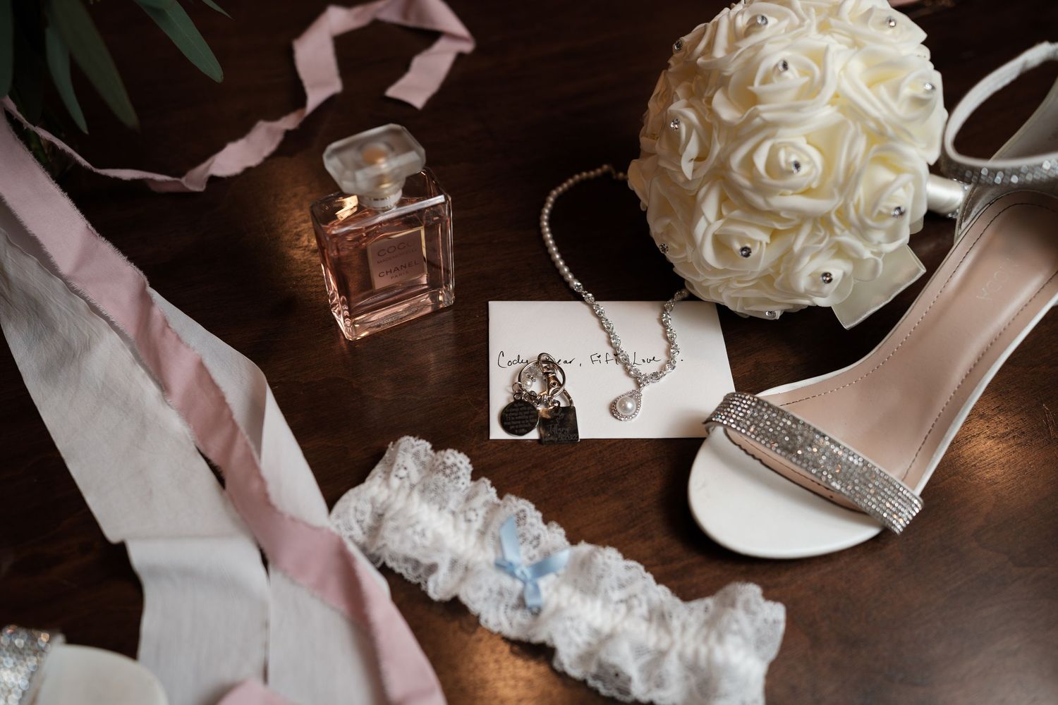 Wedding accessories including white shoes, garter, perfume bottle and delicate floral details arranged on dark wooden surface.