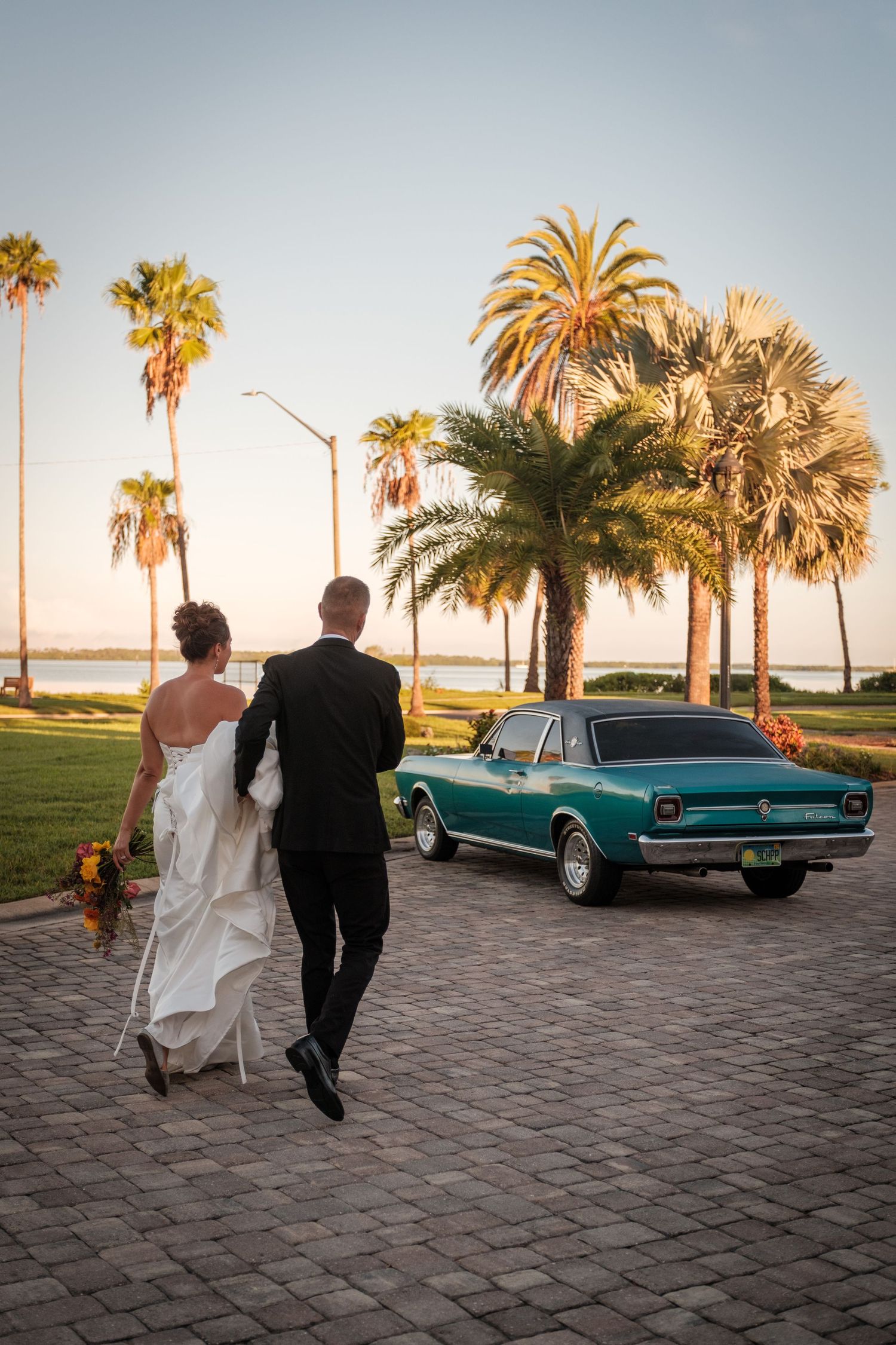 Bride and groom walk toward a vintage teal car surrounded by palm trees at sunset near a waterfront.