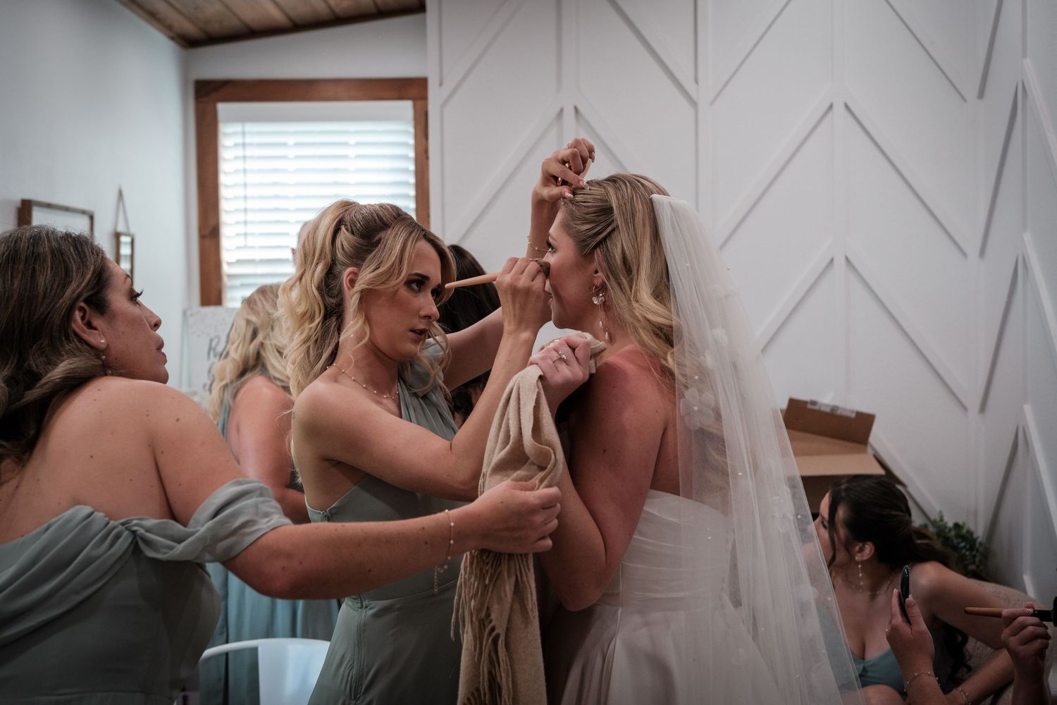 Bridesmaids in sage green dresses help bride with veil and dress in bright white room before wedding ceremony.