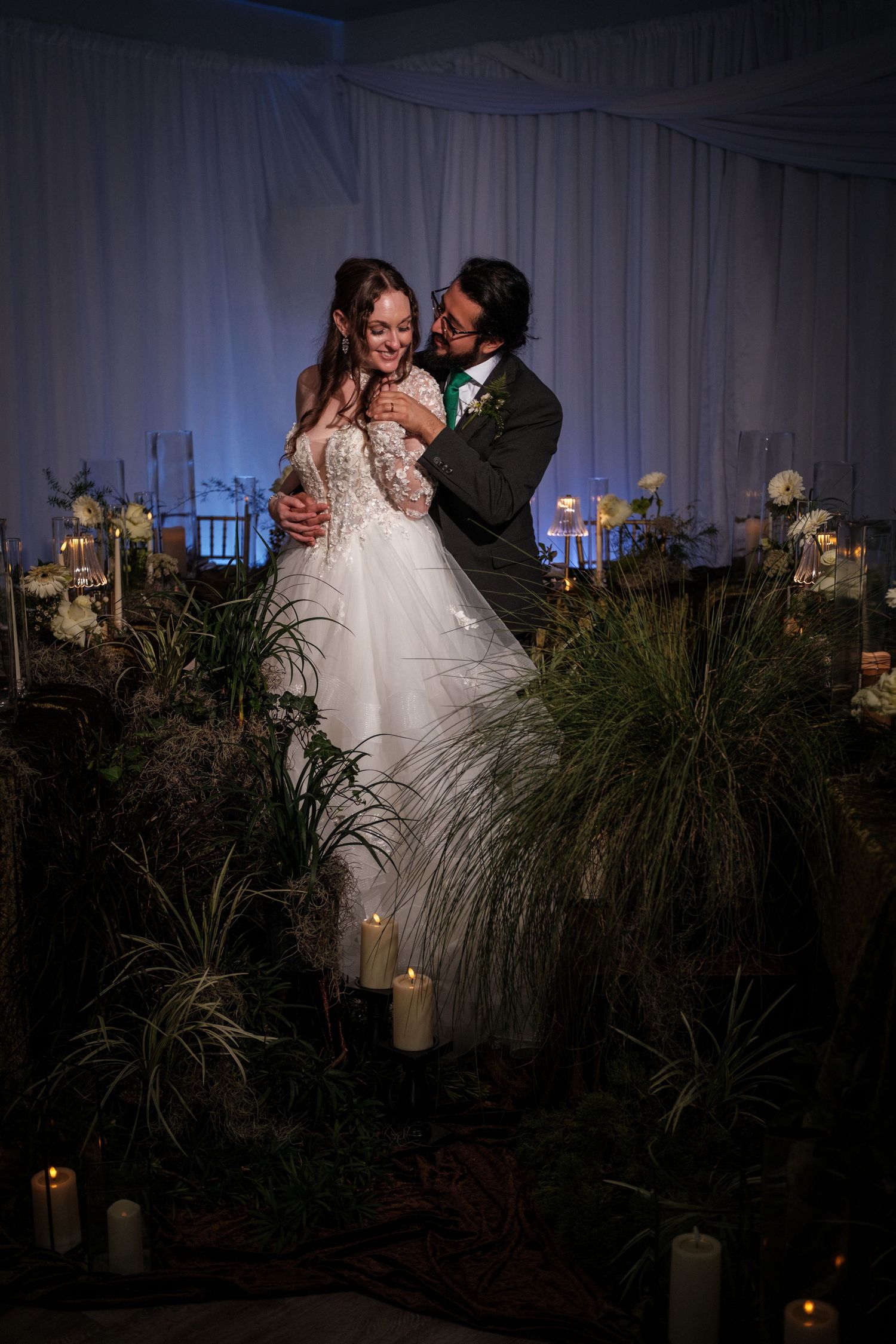 Bride and groom share a romantic embrace surrounded by candles and lush greenery at an elegant evening wedding reception.