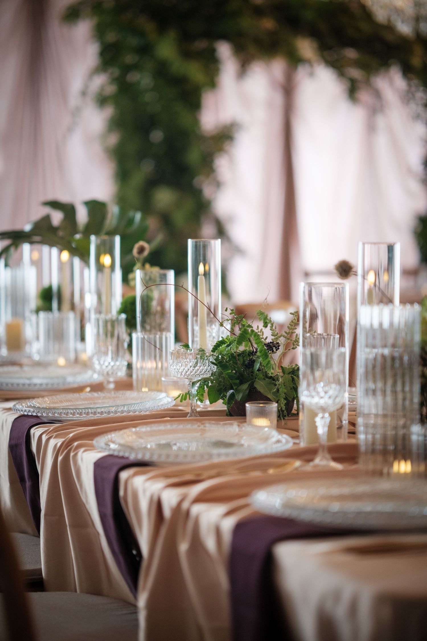 Elegant wedding reception table with purple and gold linens, crystal glassware, candles, and lush greenery centerpieces.