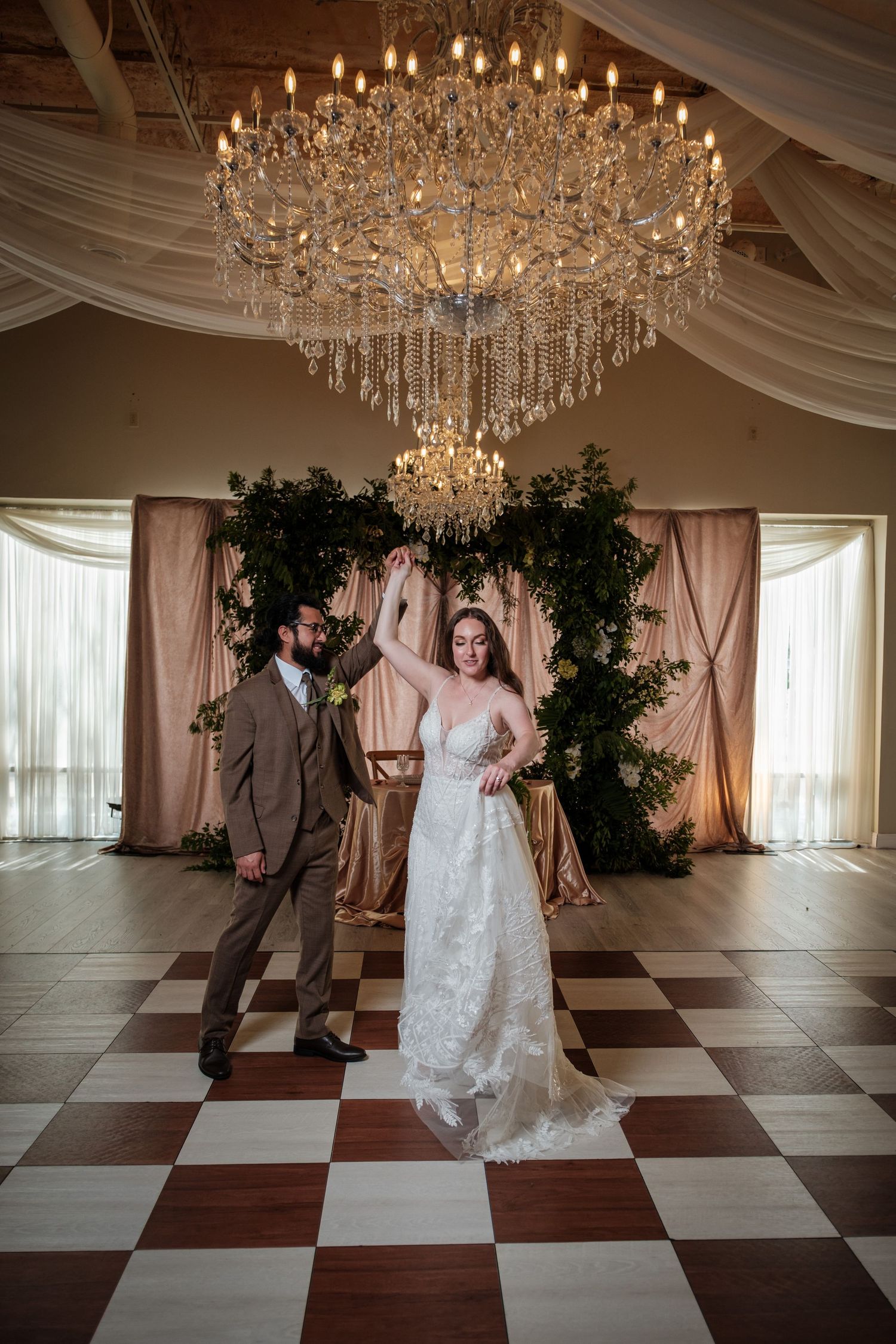 Bride and groom dancing under a grand crystal chandelier on a checkered dance floor in an elegant wedding venue.