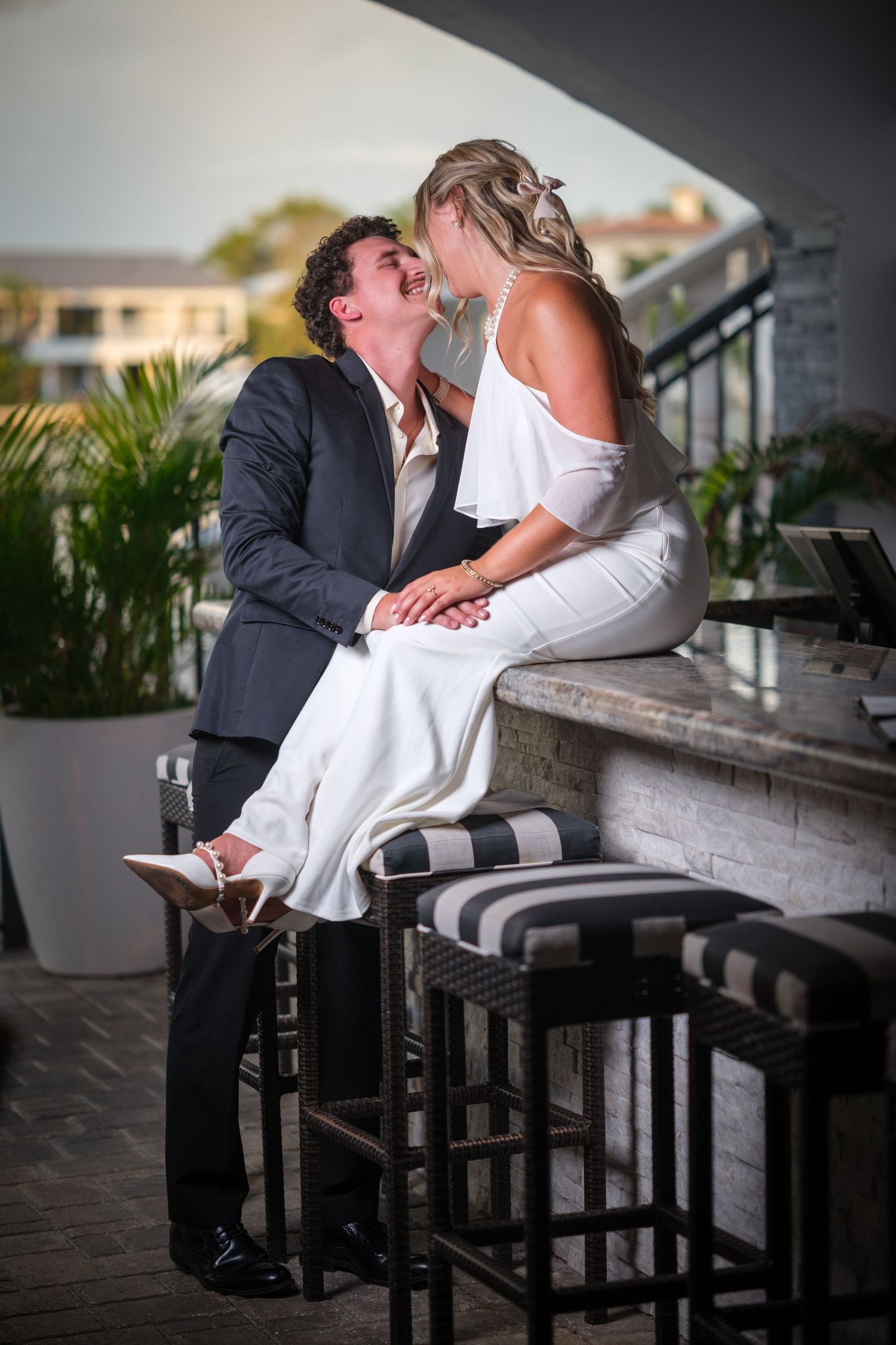 A bride in white sits on a bar counter as her groom in a dark suit leans in for a kiss on a rooftop venue.