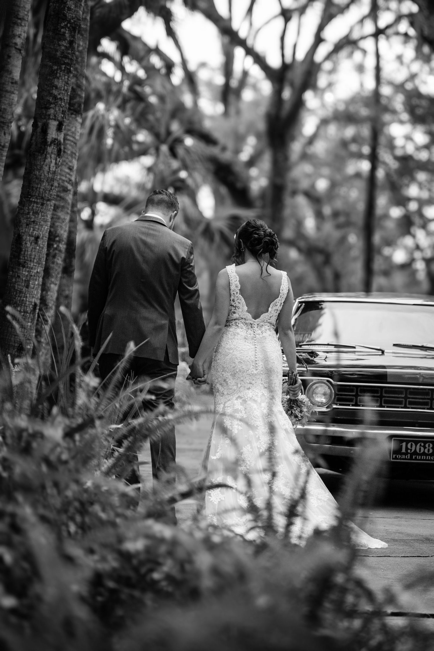 A black and white photo of a couple in wedding attire walking beside a vintage car in a wooded setting.