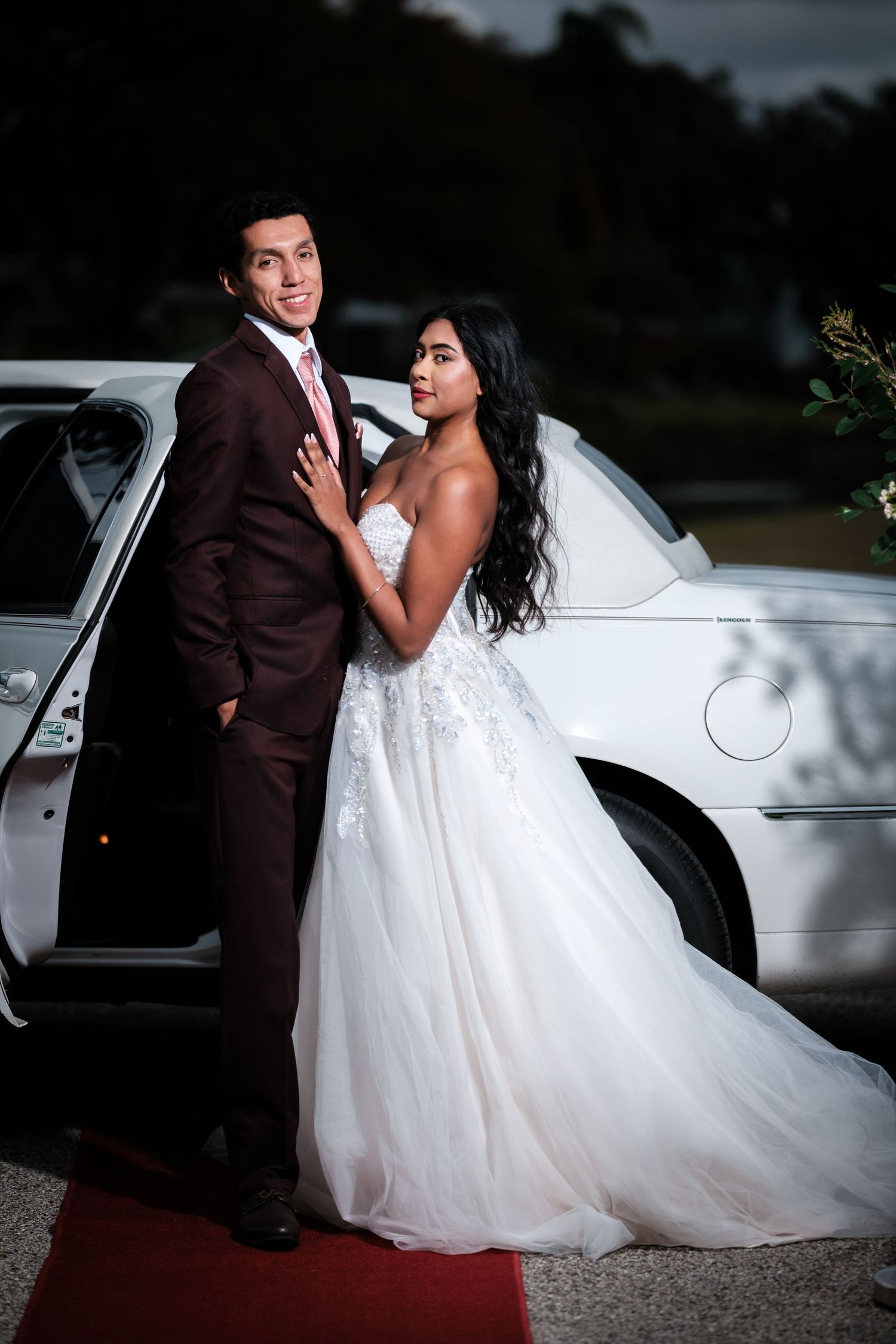 A romantic wedding photo shoot featuring a couple posing by a classic white car at dusk.