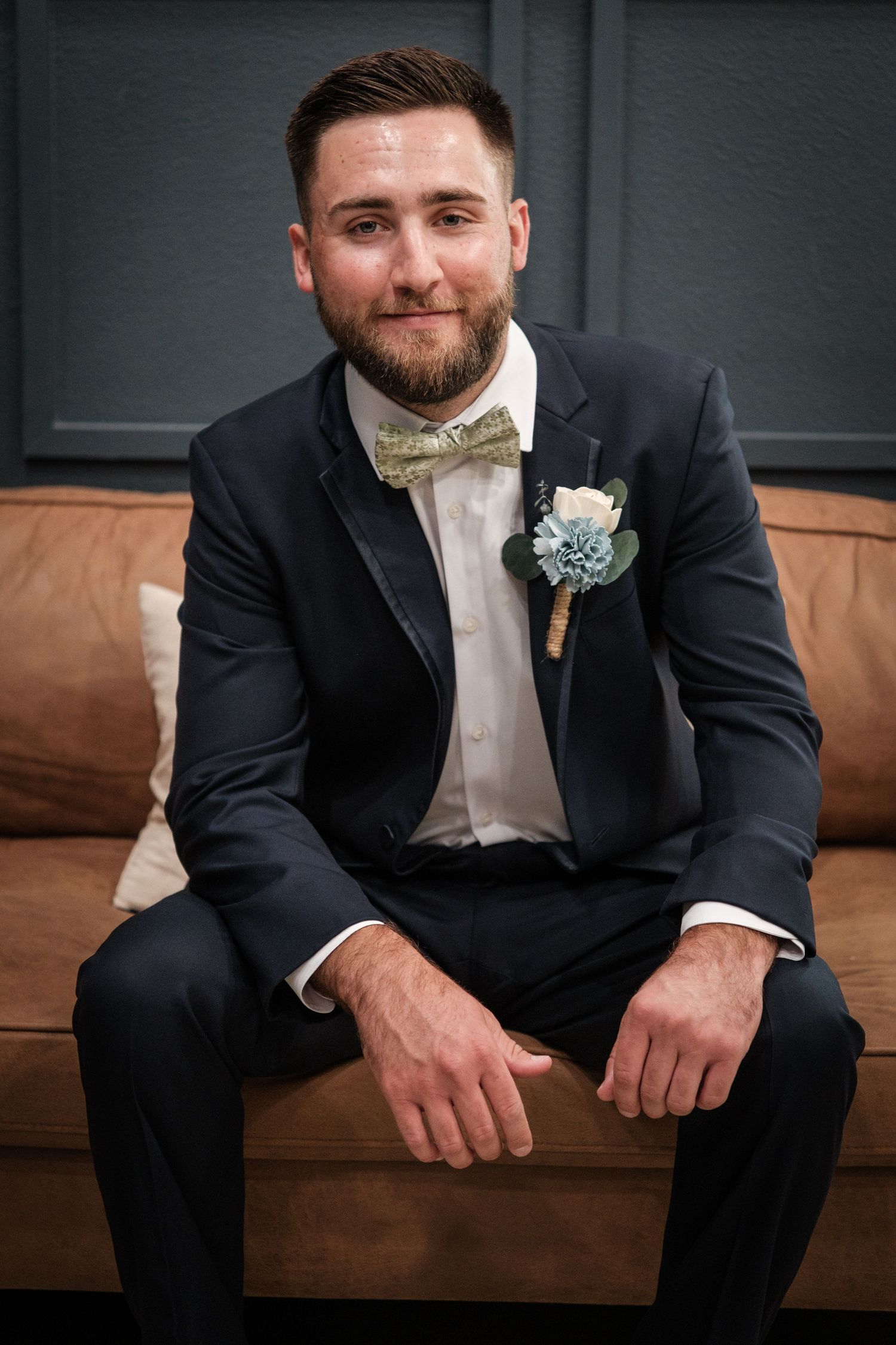 Groom in navy suit with floral boutonniere and gold bow tie seated on leather sofa smiling at camera.