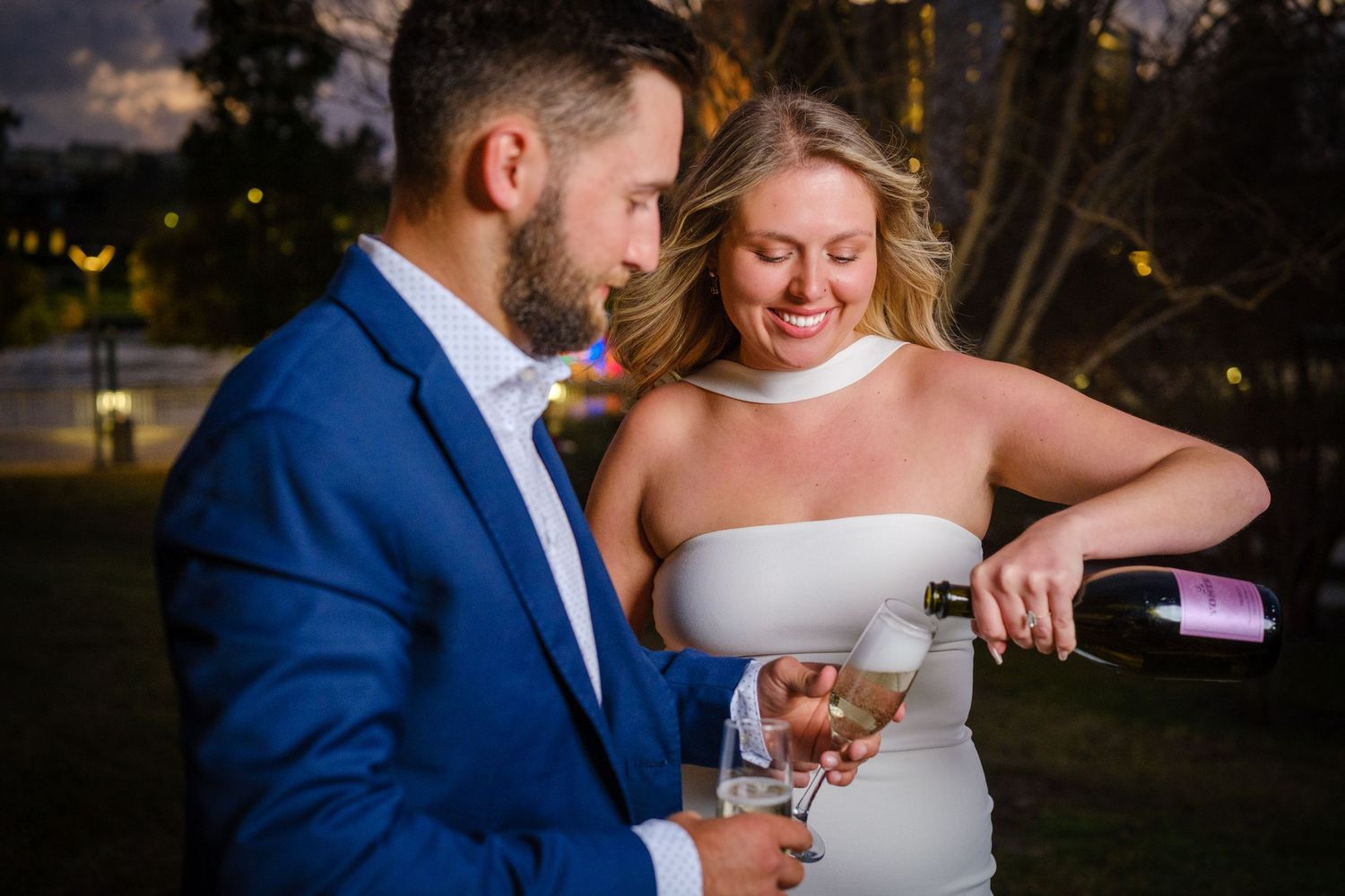 Couple in elegant attire pouring champagne outdoors at dusk, woman smiling in white strapless dress.
