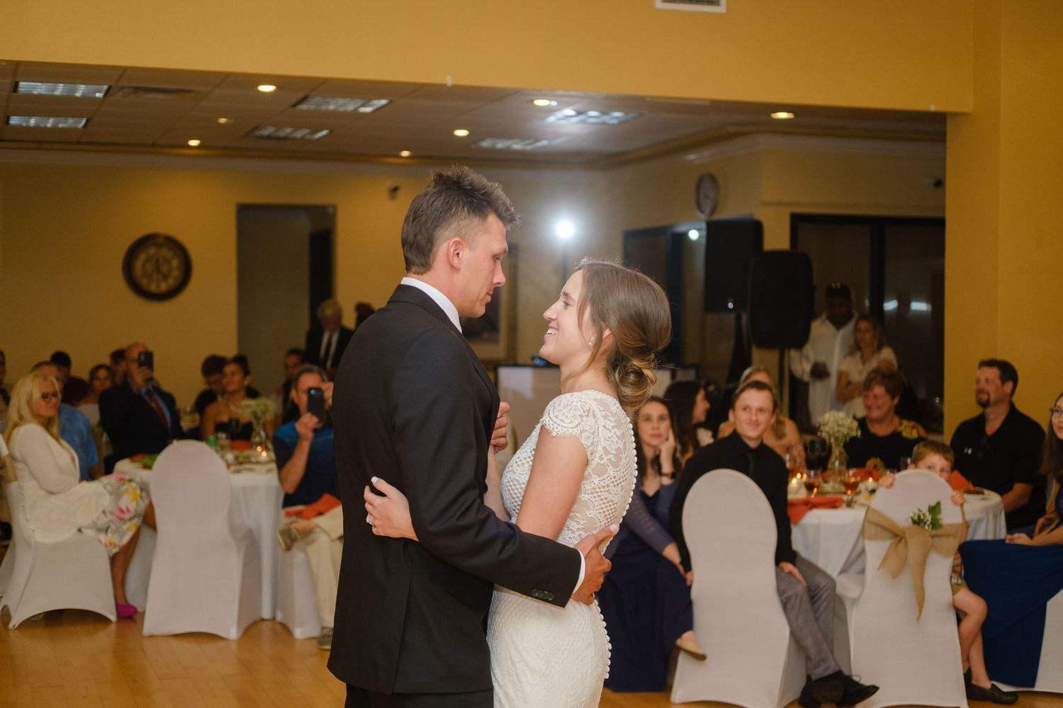 Wedding guests dance and celebrate together in a reception hall decorated with white chair covers and warm lighting.