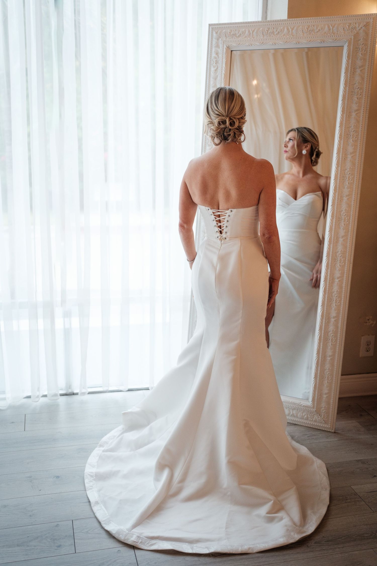 Bride in a strapless white wedding gown with lace-up back and train, viewing her reflection in an ornate white mirror.