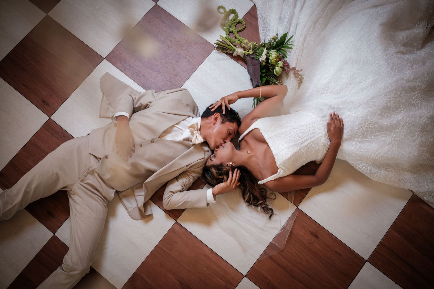 Bride and groom lying on checkered floor sharing a romantic kiss, with bridal bouquet nearby.