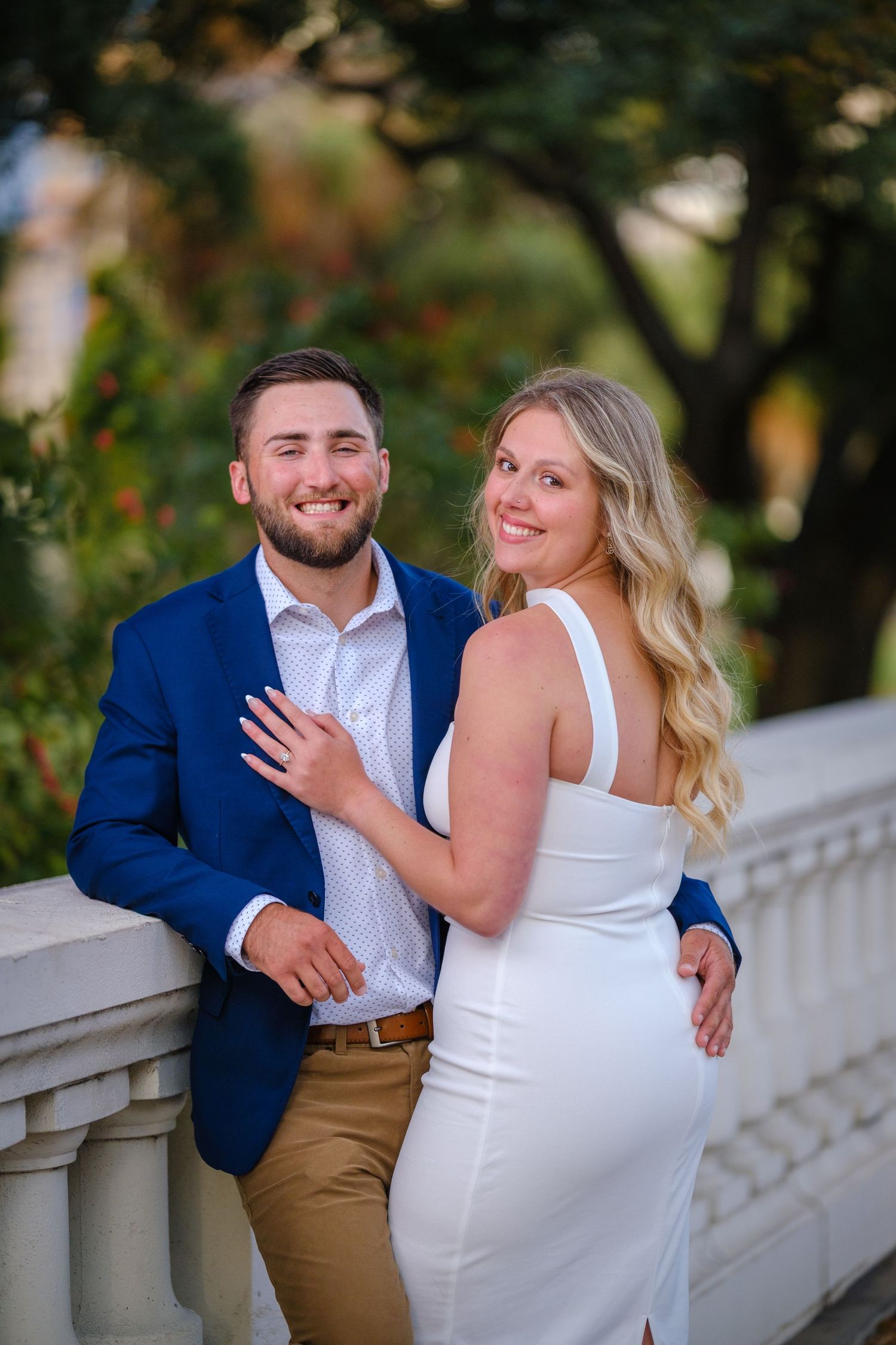 Smiling couple in white dress and blue blazer posing by stone railing with lush greenery backdrop.