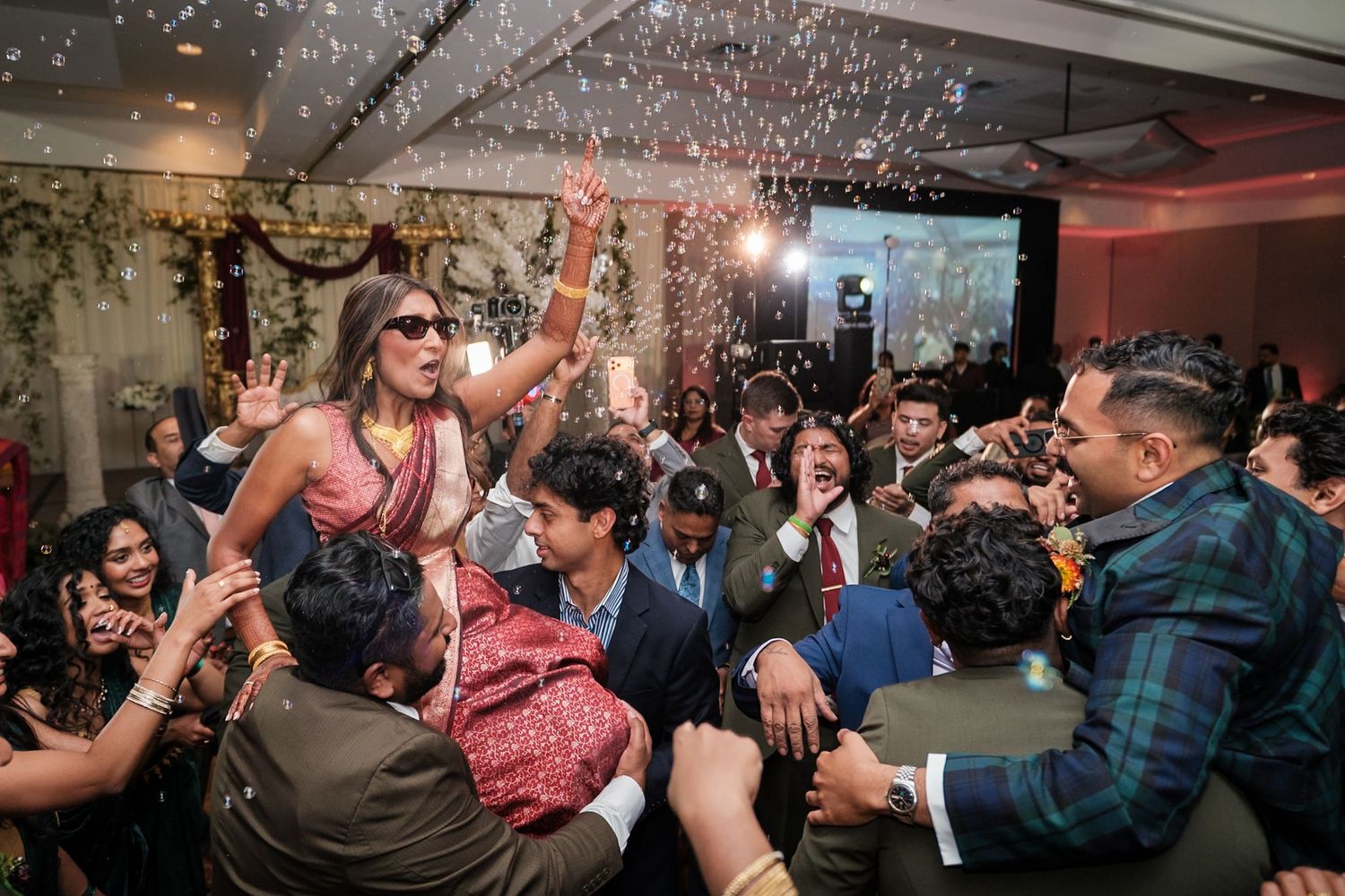 Energetic crowd celebrates at a party with confetti and dancing as someone is lifted up in excitement on the dance floor.