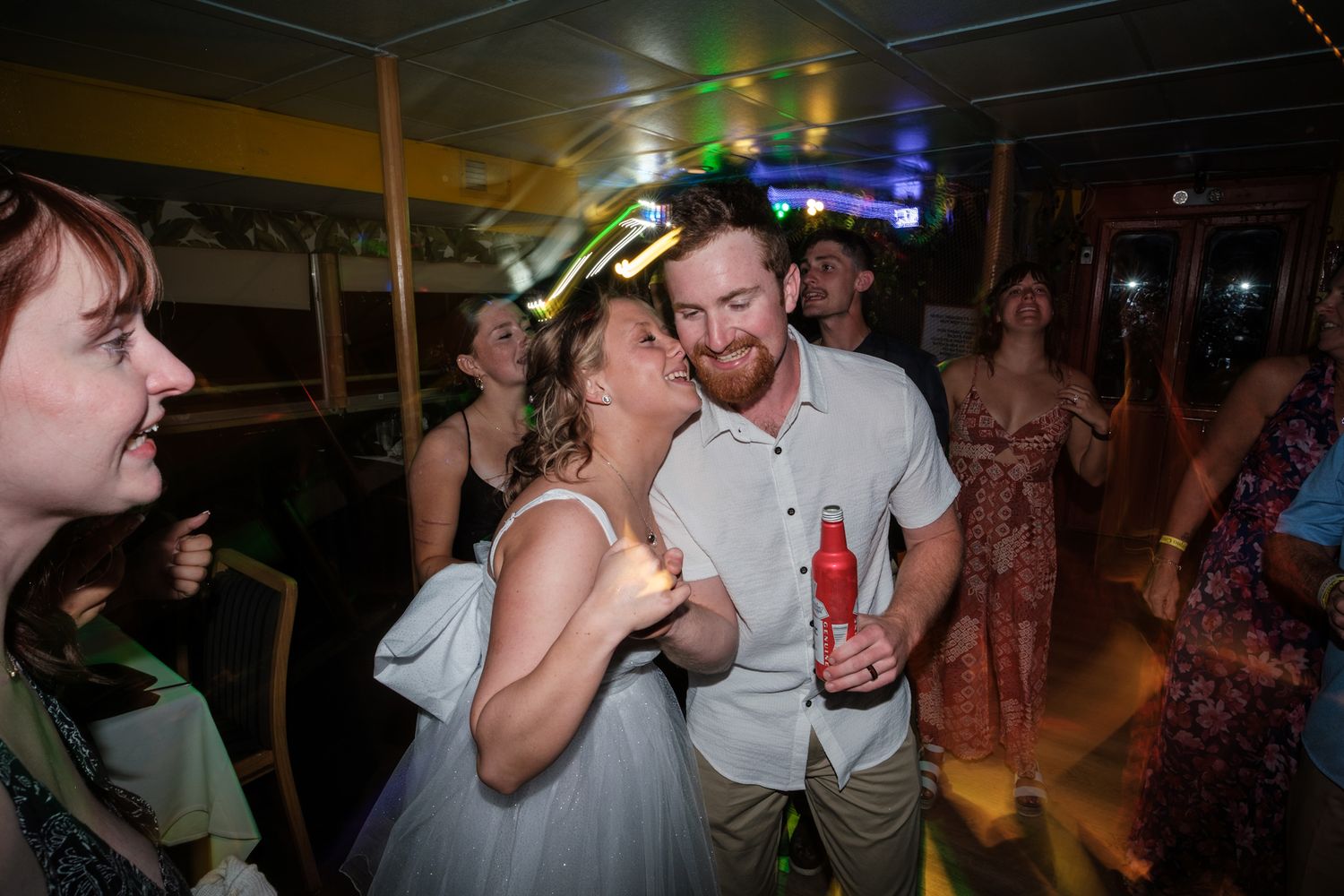 Wedding guests dance and celebrate on a dimly lit dance floor with colorful party lights.