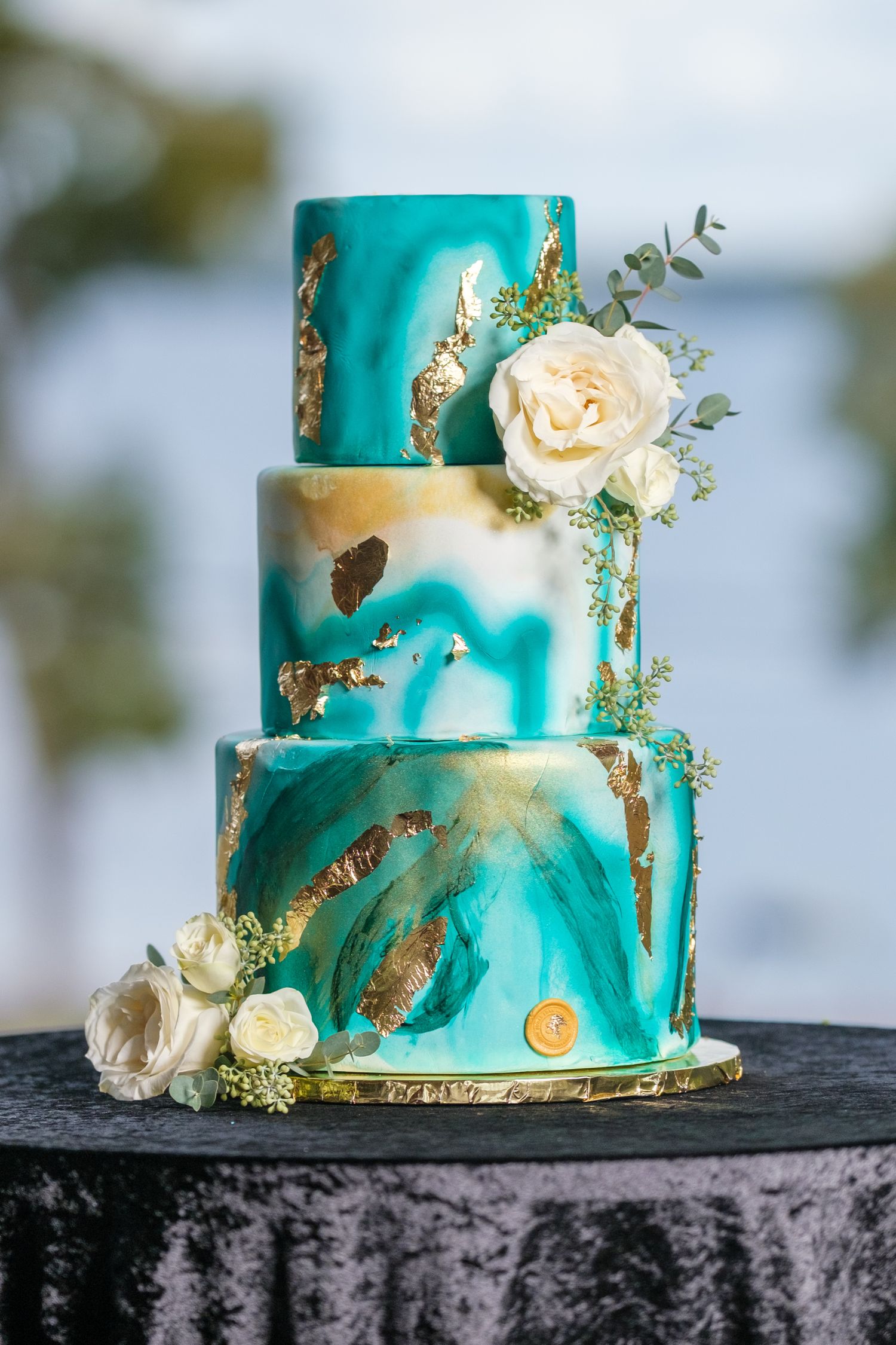 Elegant turquoise wedding cake with gold leaf accents and fresh cream roses displayed on a dark stone surface.
