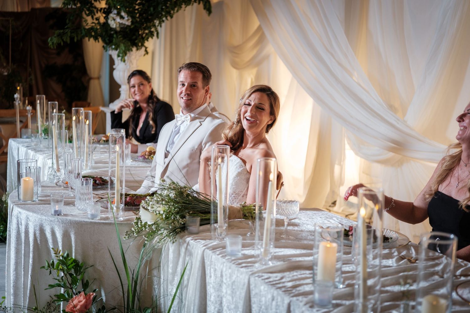 Bride and groom smiling joyfully at elegant wedding reception head table with candles and white drapery backdrop.