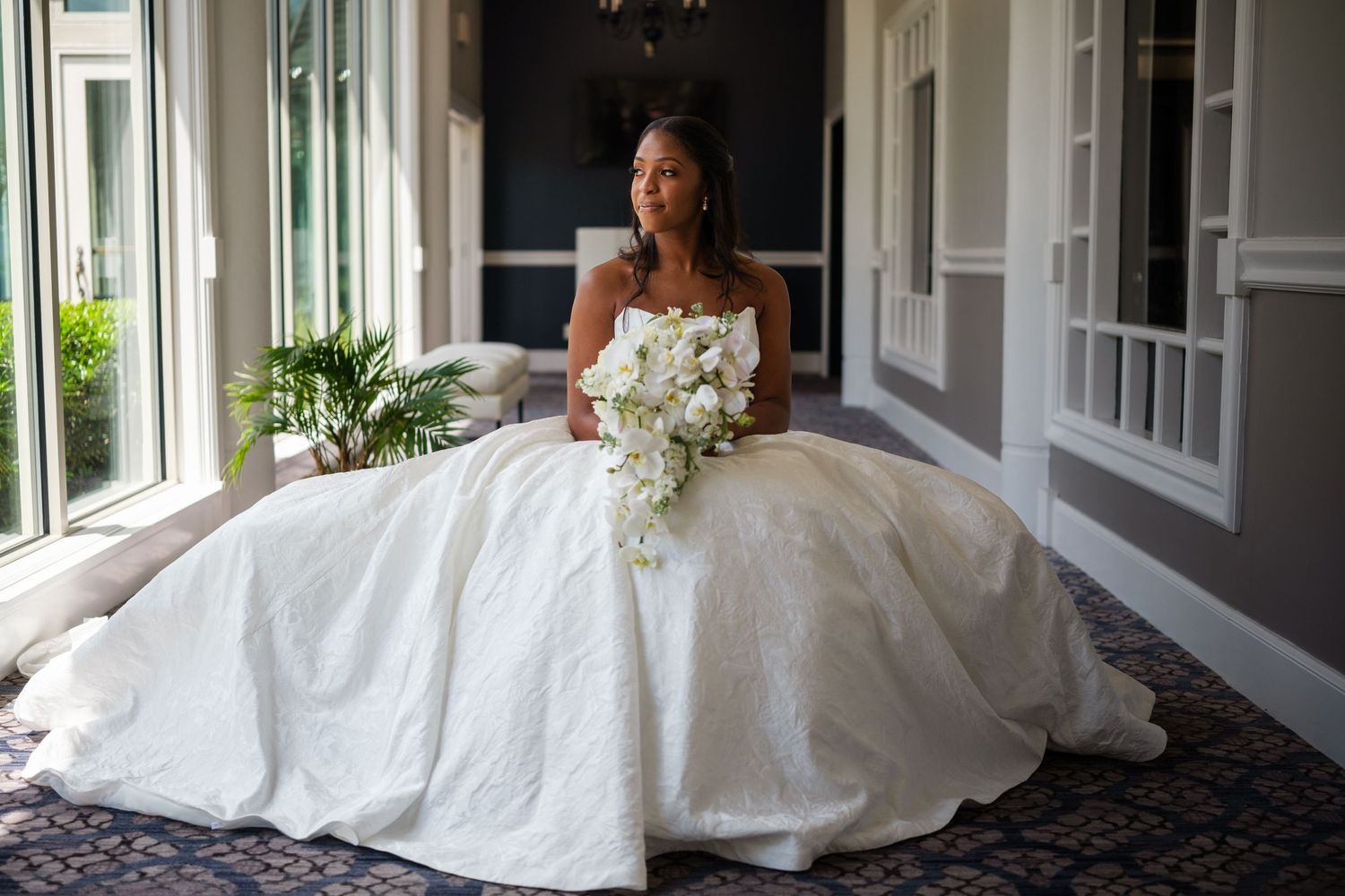 Bride in a strapless white ball gown holding a white floral bouquet, seated in an elegant venue hallway.