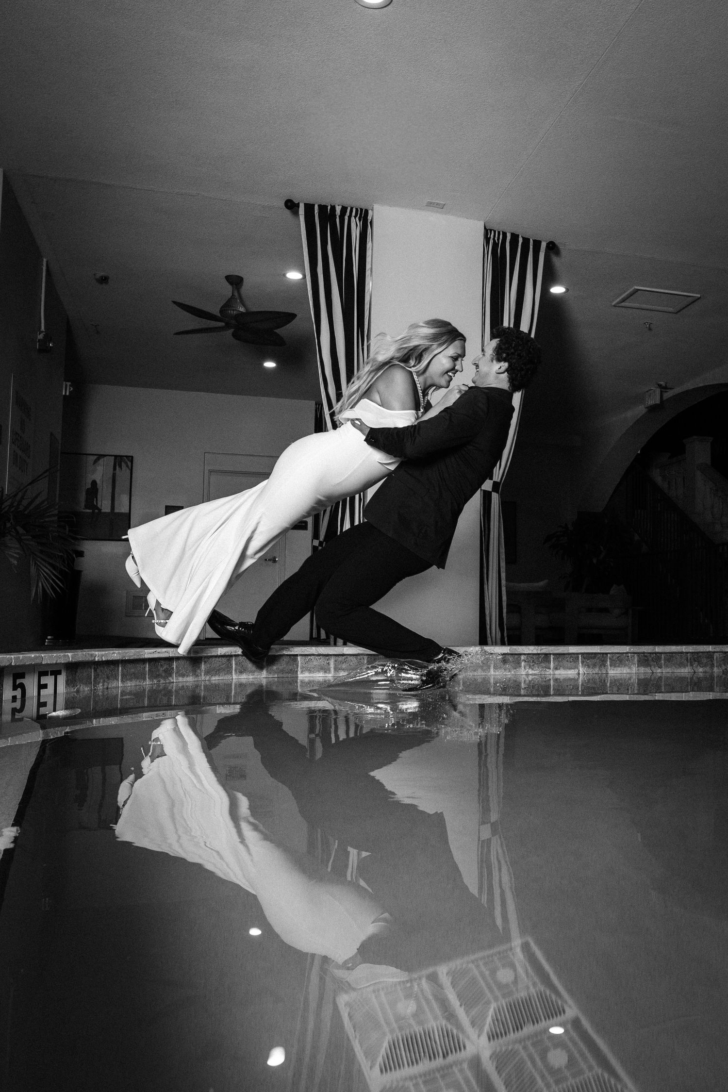 Black and white photo of a couple in wedding attire sharing a romantic dip kiss beside a reflective pool indoors.