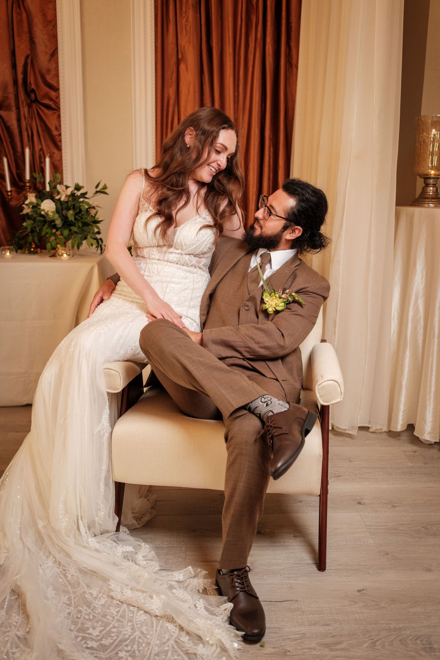 Bride sitting on groom's lap in a white lace gown as he wears a brown suit with boutonniere in elegant venue.