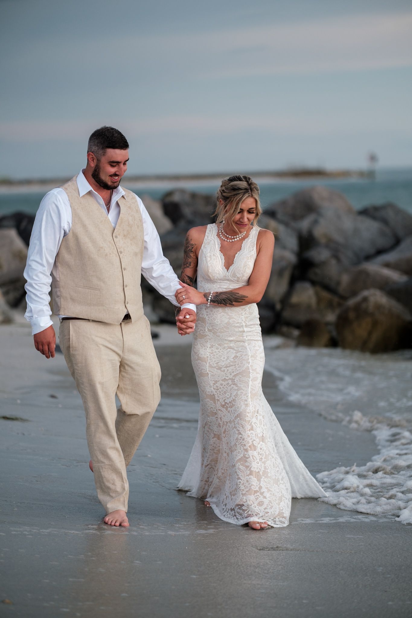 Newlywed couple walking barefoot along the beach shoreline at sunset in matching beige and white attire.