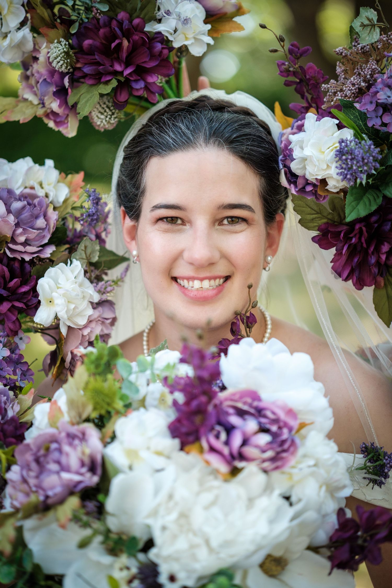 Smiling bride in white veil surrounded by purple and white floral arrangement holding bouquet of peonies outdoors.