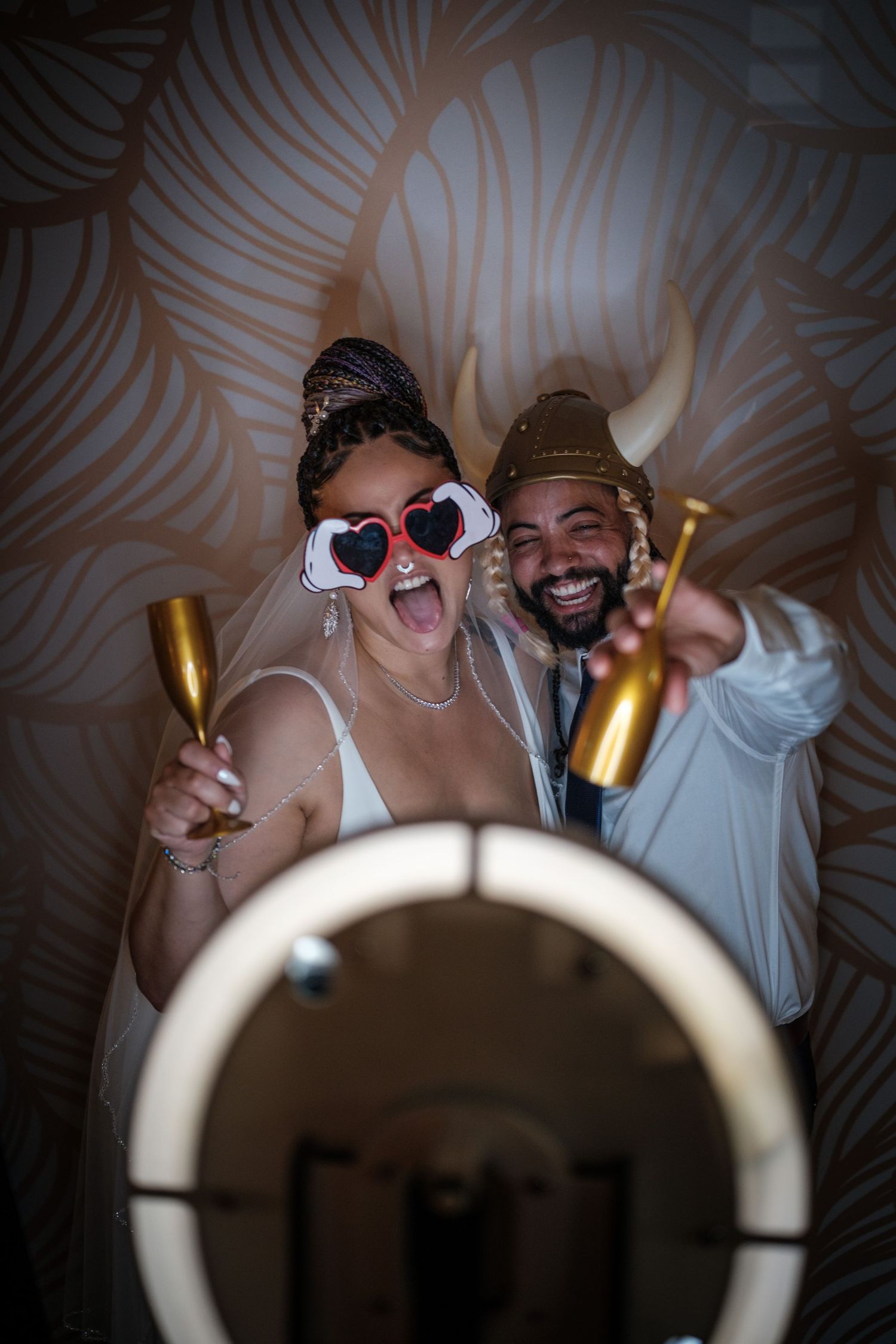 Joyful couple in wedding attire posing in photo booth with heart sunglasses, gold crown, and champagne bottles.
