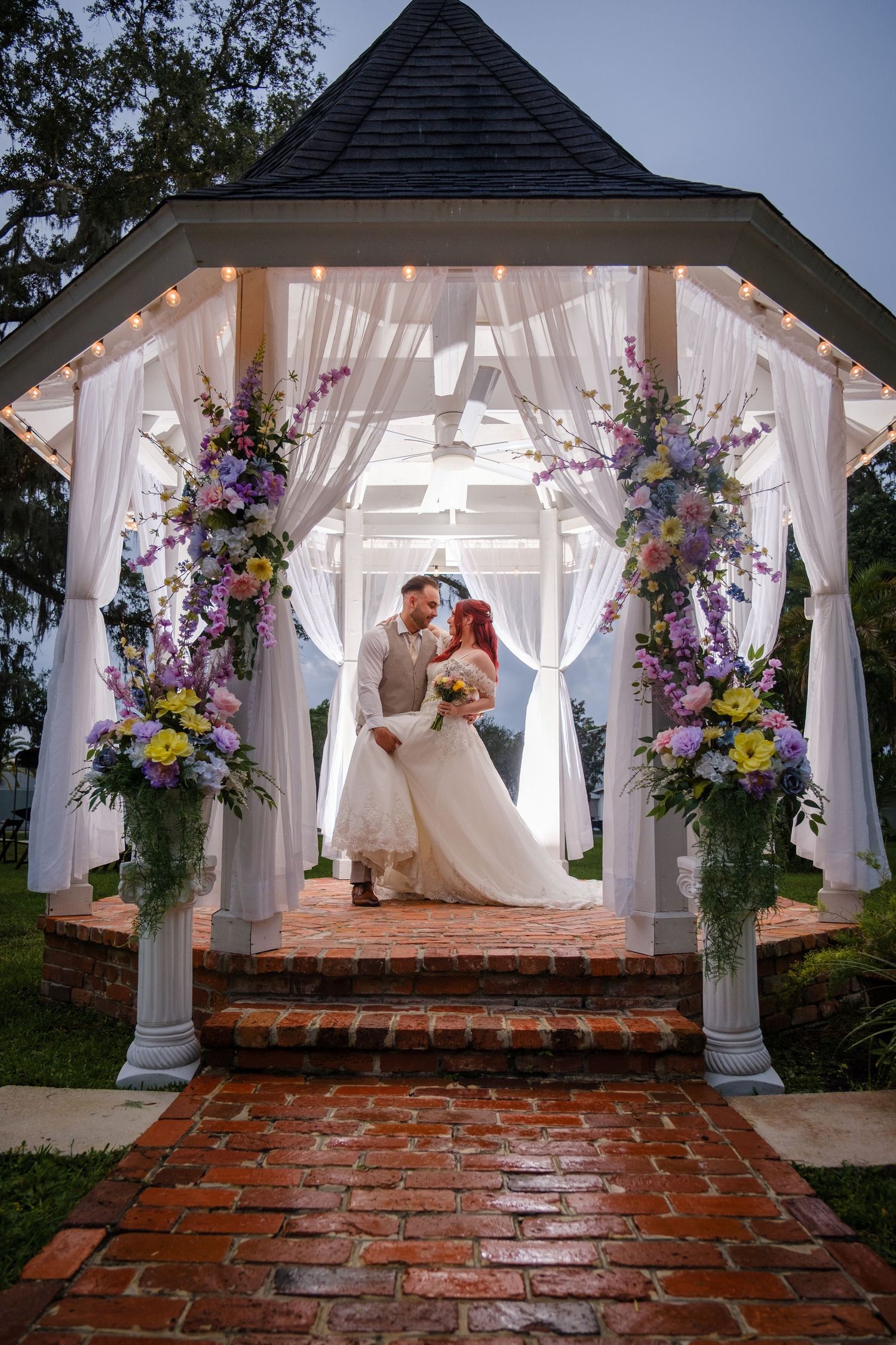 Wedding ceremony gazebo decorated with white drapes and purple floral arrangements on a brick path at sunset.