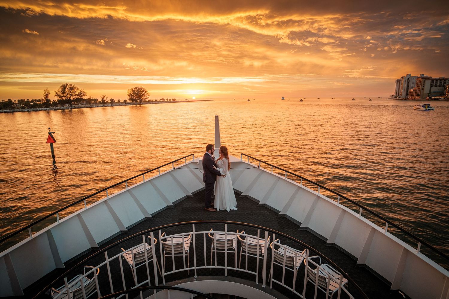 Couple embracing on ship bow at golden sunset over calm water, romantic wedding photo with vibrant orange sky.