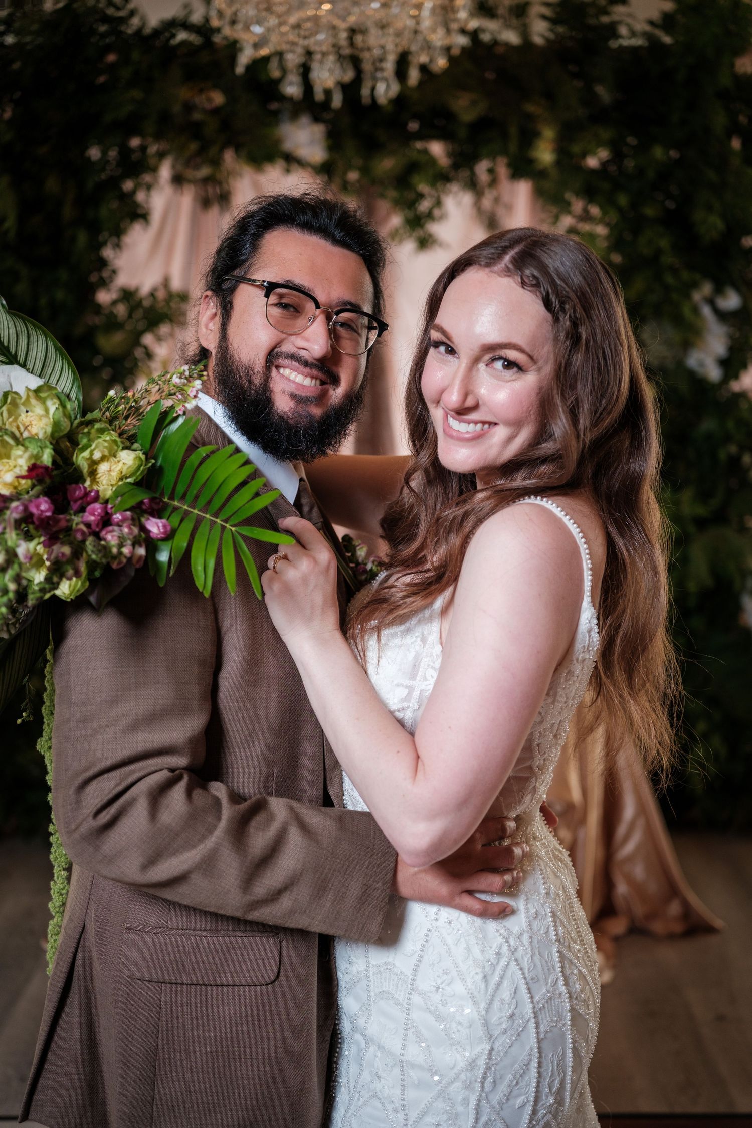 Smiling bride and groom pose together at wedding venue with lush greenery and floral bouquet backdrop.