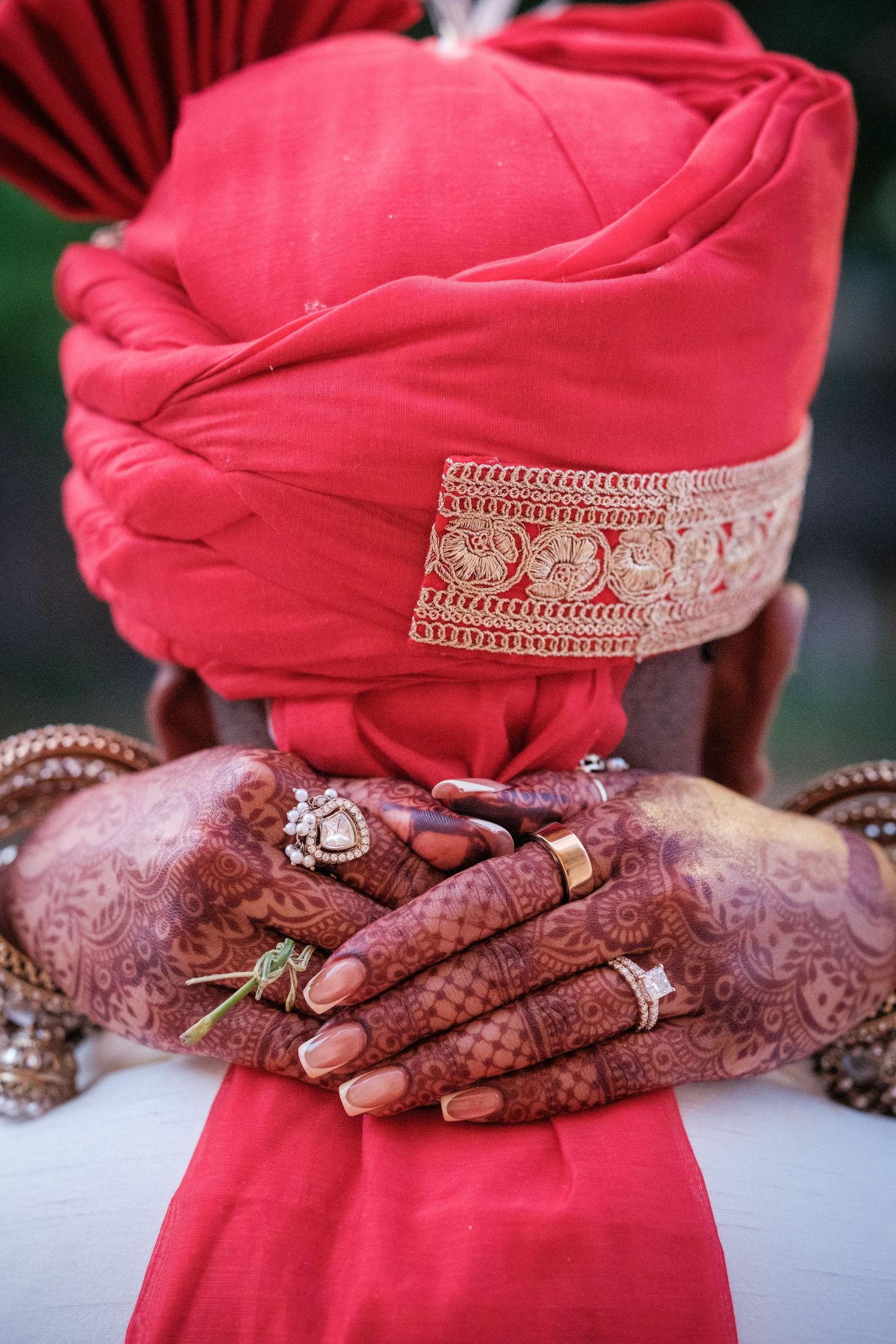 Indian groom in red turban with gold trim, hands adorned with henna mehndi and rings at wedding ceremony.