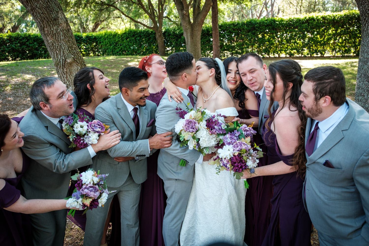Wedding party kissing and celebrating outdoors, bridesmaids in purple dresses and groomsmen in gray suits.