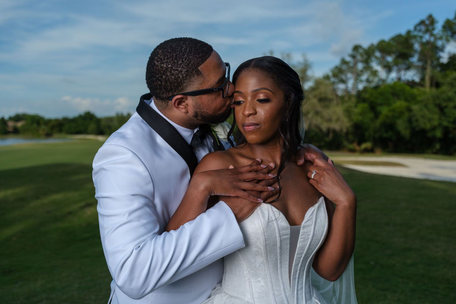 A groom in a white suit embraces his bride in a strapless wedding gown outdoors on a lush green golf course.